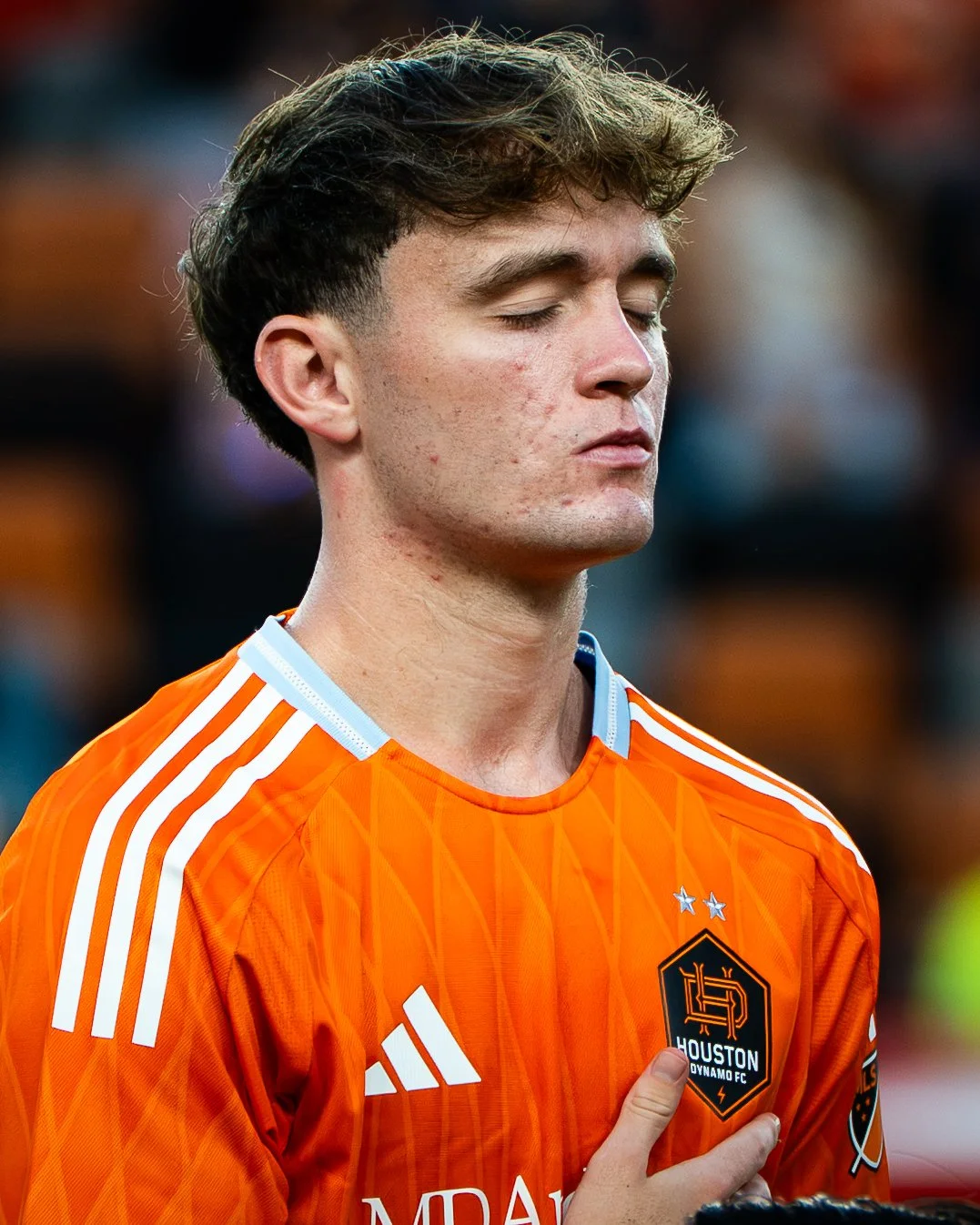 A young man with closed eyes and sweaty face wearing an orange Houston Dynamo FC soccer jersey with the team crest near his chest, standing with his hand over his heart.