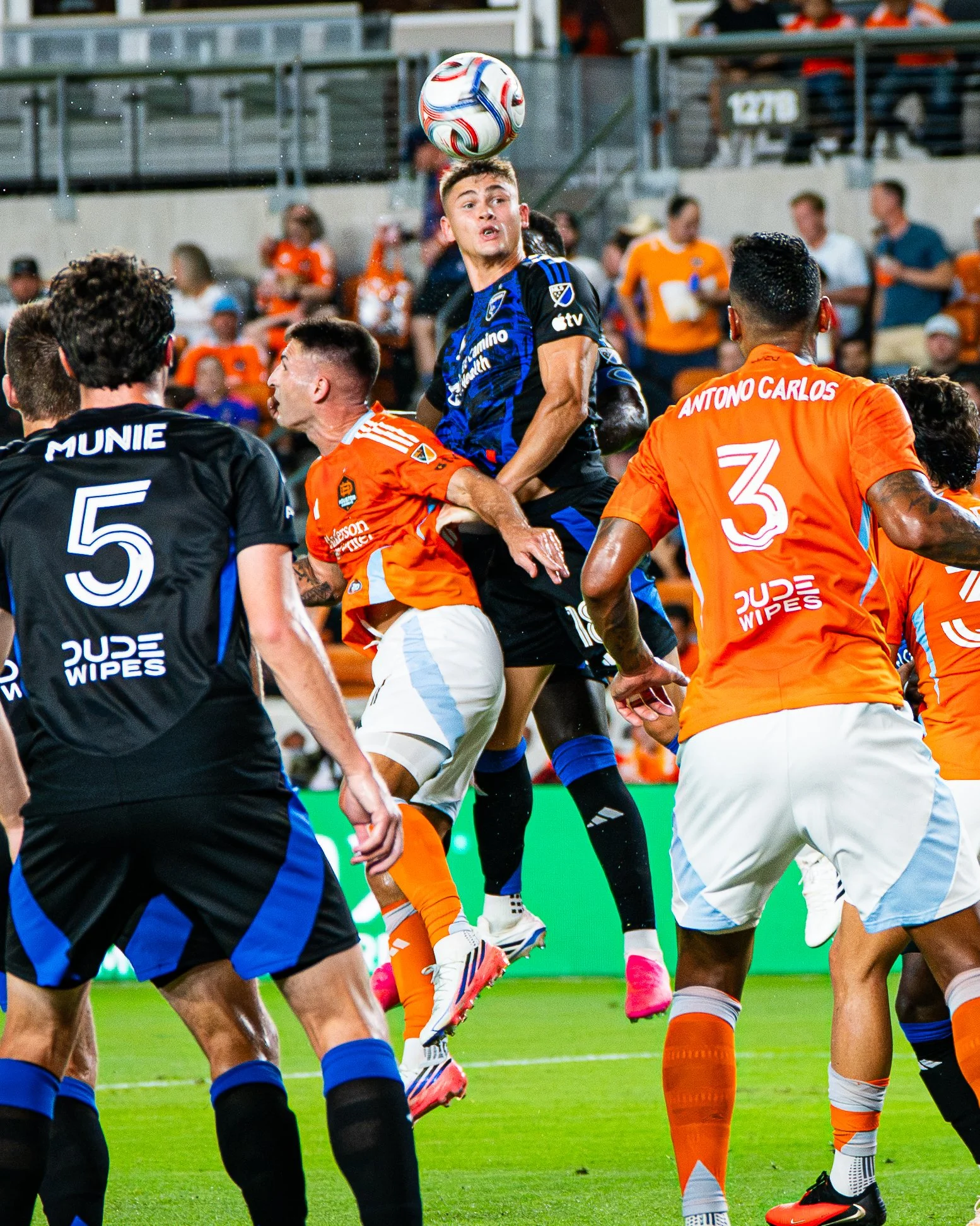 A soccer player in a blue jersey jumps to head the ball during a match, surrounded by players in orange and black jerseys on a green field with spectators in the background.