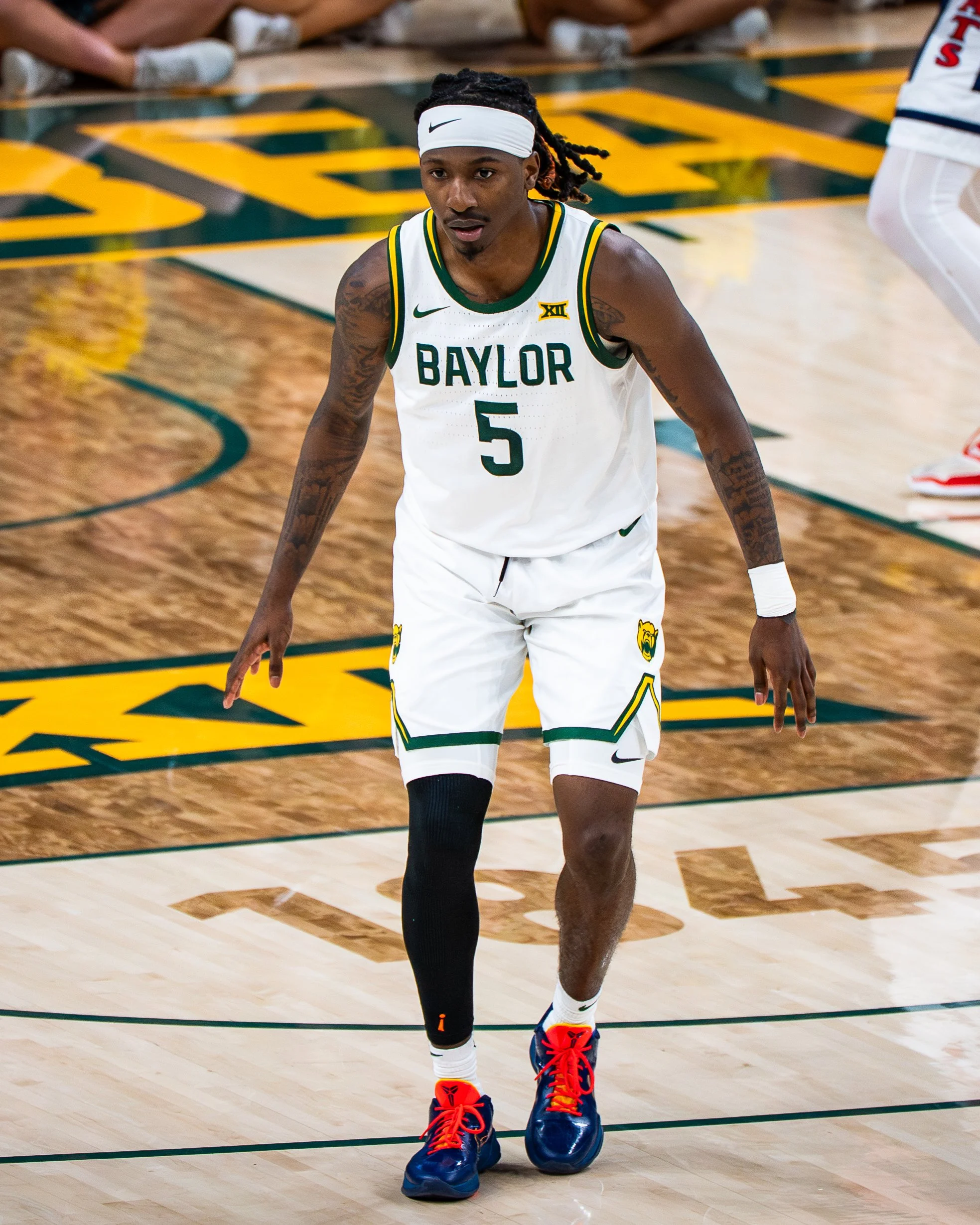 An African American basketball player wearing a white Baylor jersey with the number 5 and a white headband on a basketball court. The player has dreadlocks and tattoos and is standing in a defensive stance.