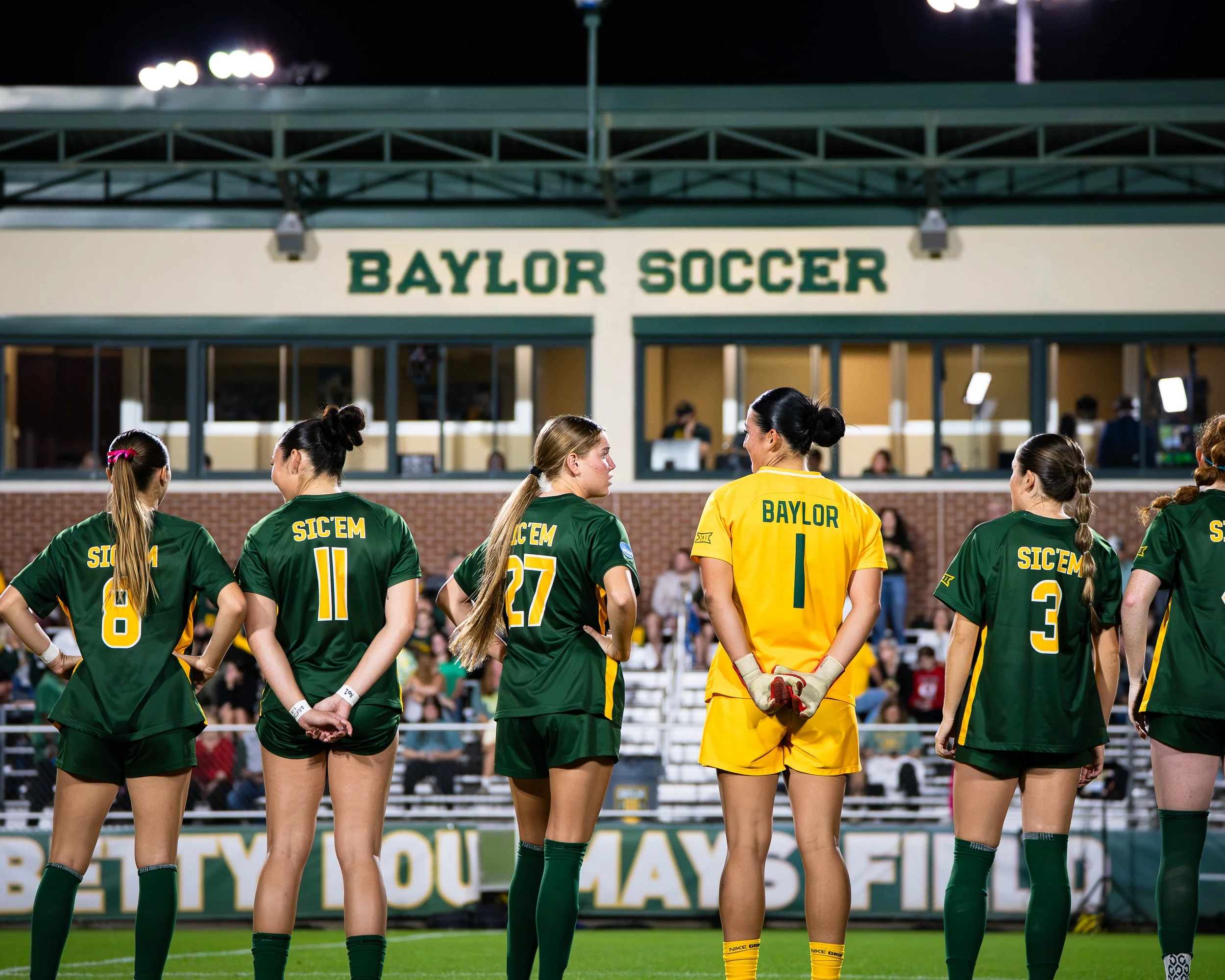 Women soccer players from Baylor team standing on the field during a game at night, with the stadium and crowd in the background, some players have their hands behind their backs and are facing the coach or referee.