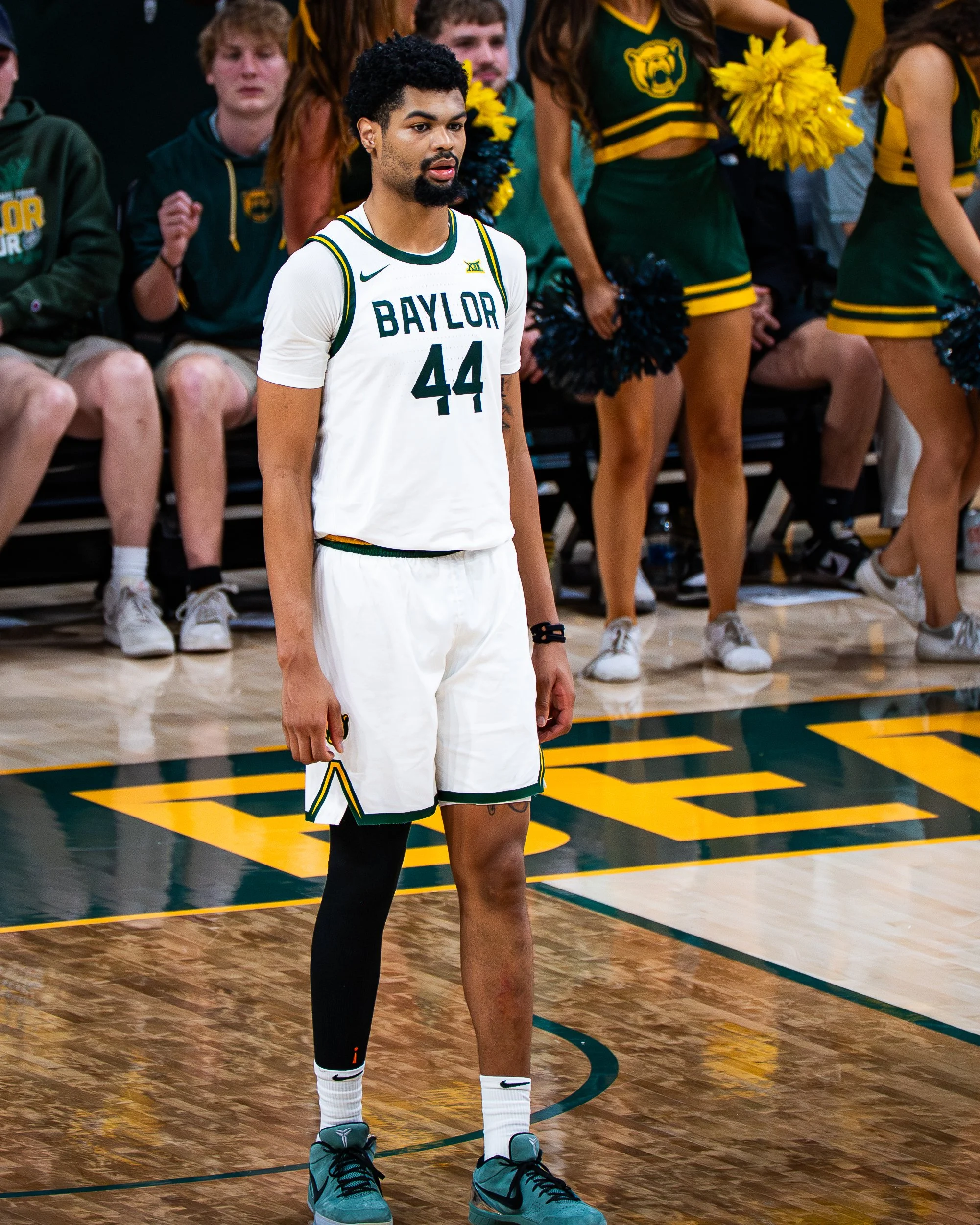 A basketball player from Baylor University wearing a white jersey with the number 44, standing on a basketball court, with cheerleaders and spectators in the background.