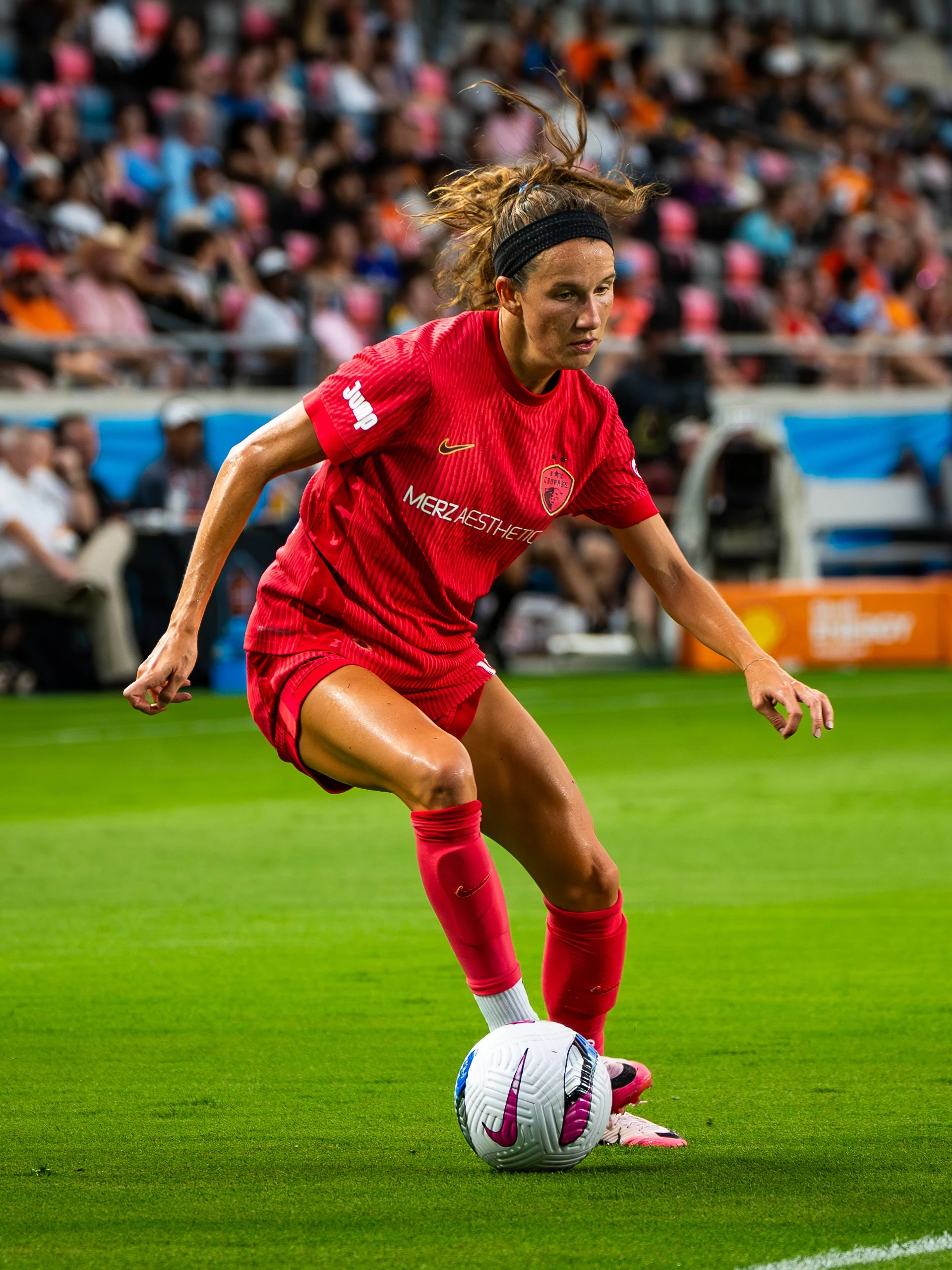 A female soccer player in a red jersey is about to kick a ball during a match, with a crowd of spectators in the background.