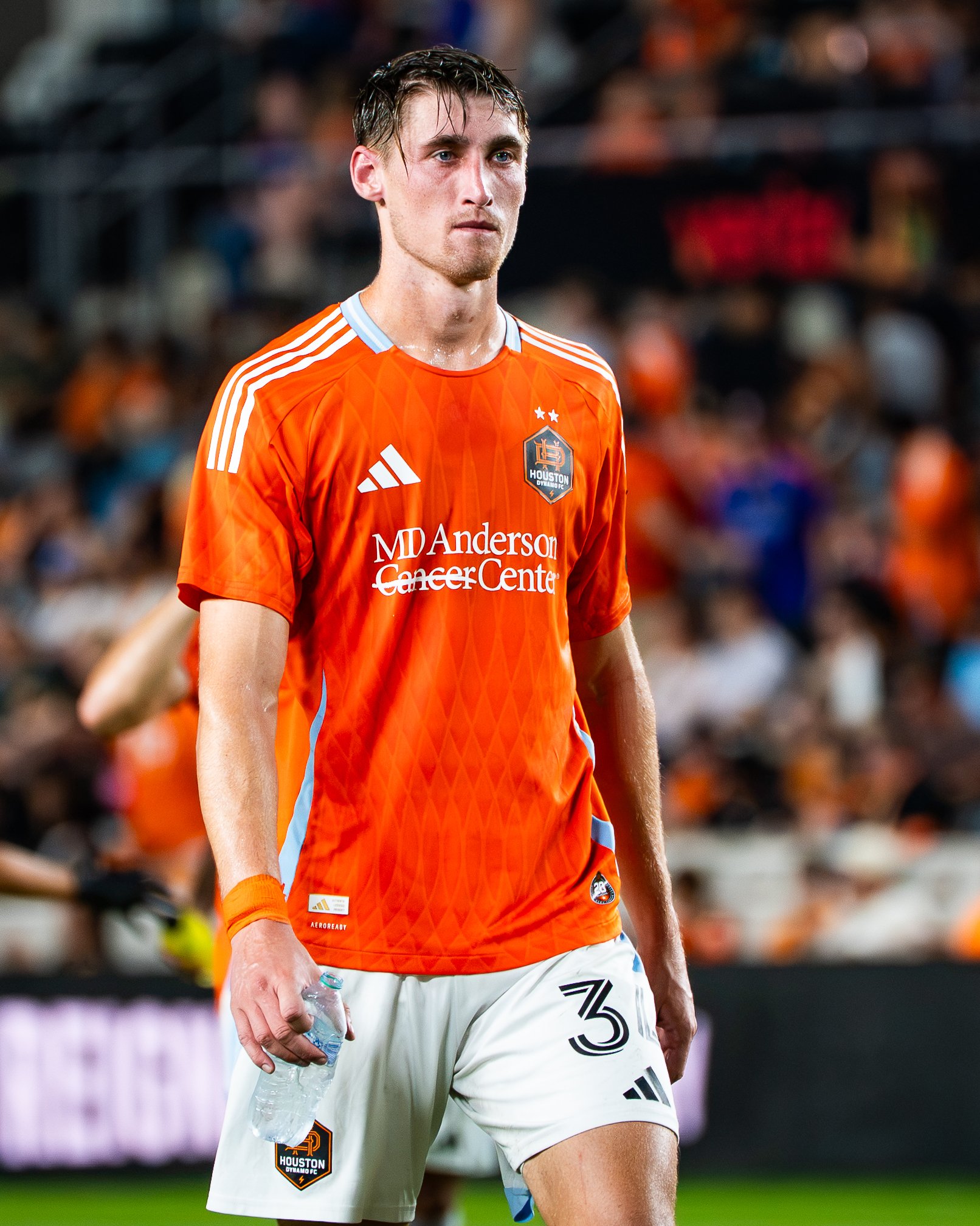 A male soccer player in an orange Houston Dynamo FC jersey holding a water bottle on a soccer field with a crowd in the background.