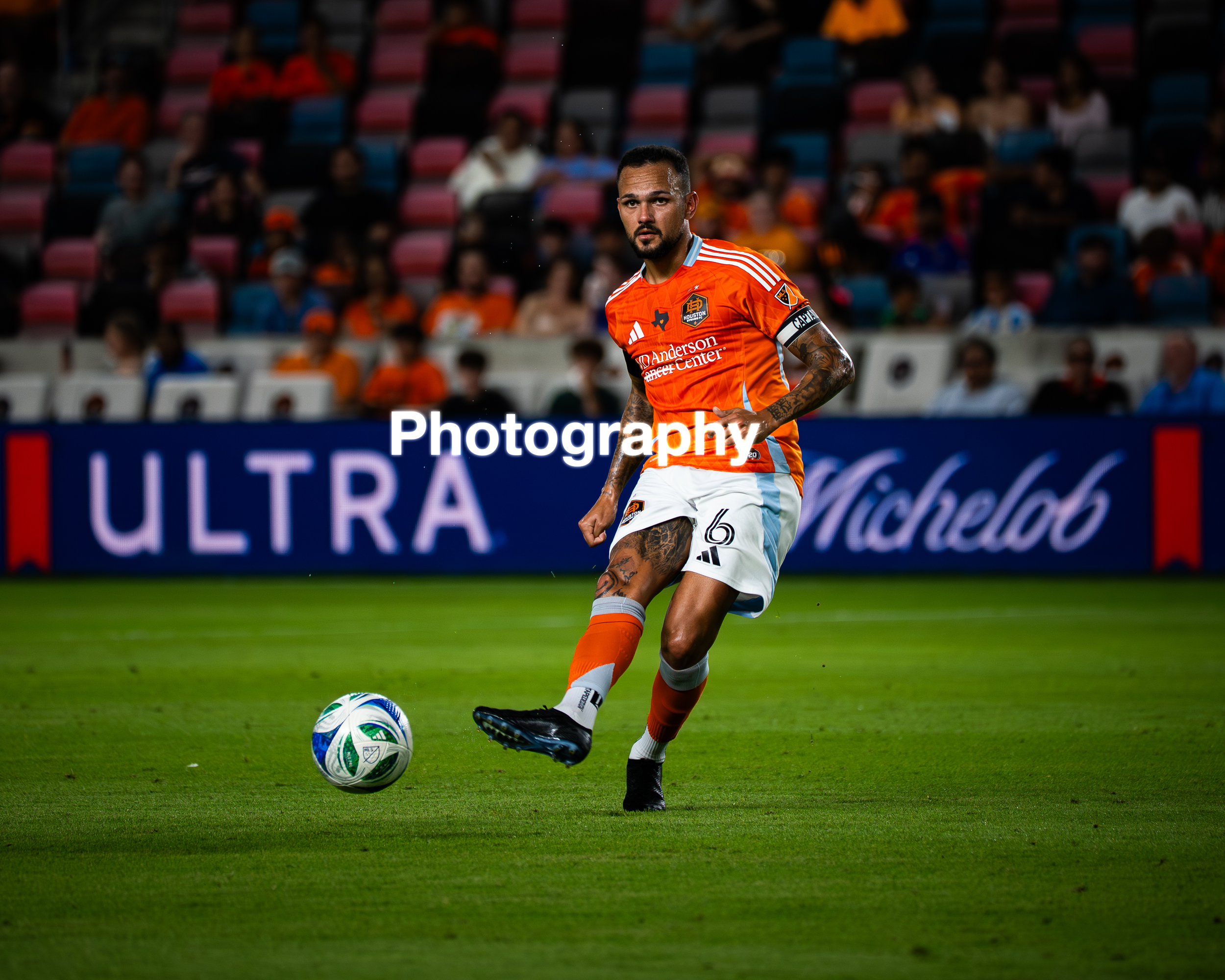 A soccer player dressed in an orange uniform with white and blue accents, wearing a captain's armband, is in mid-action kicking a soccer ball on a field during a match, with a blurred crowd in the stands in the background.