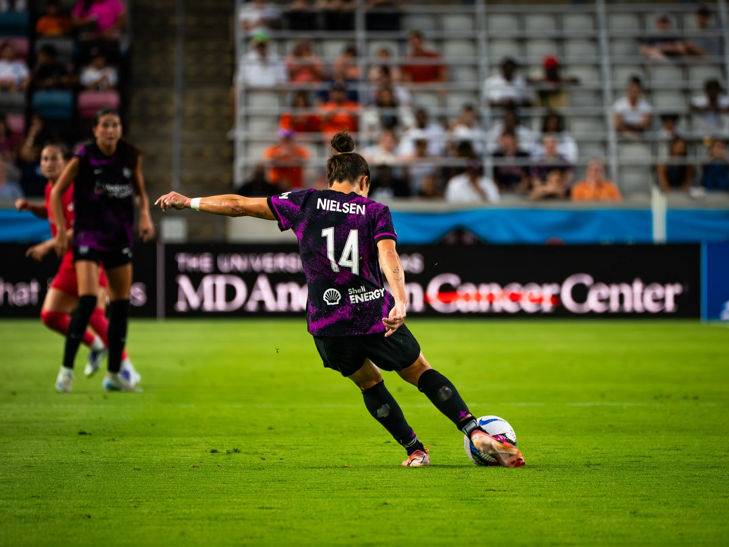 A woman wearing a black and purple soccer uniform with the name Nielsen and the number 14 on the back, kicking a soccer ball on a green field during a match. She is wearing knee braces and cleats. There are spectators in the stands in the background.