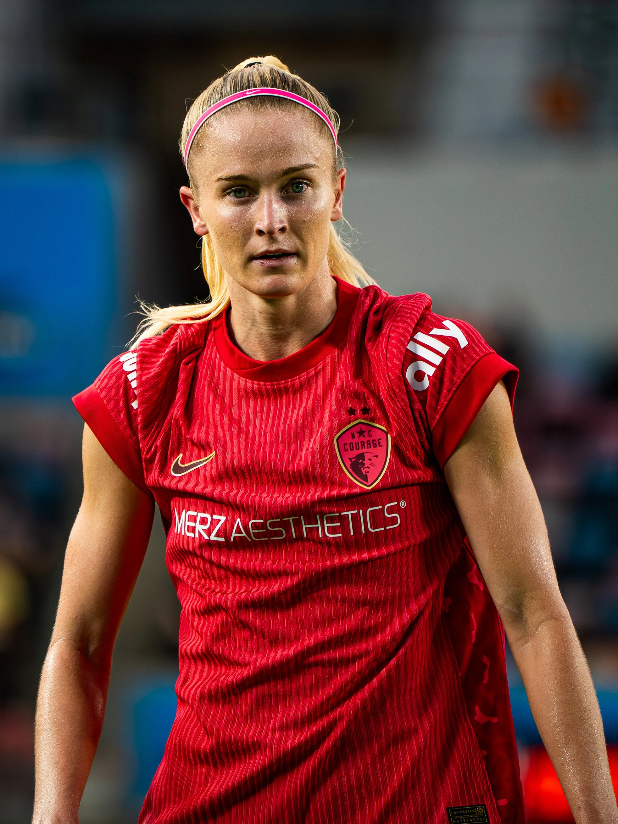 Close-up of a female soccer player with blonde hair wearing a pink headband and a red jersey with team logo and sponsor names.