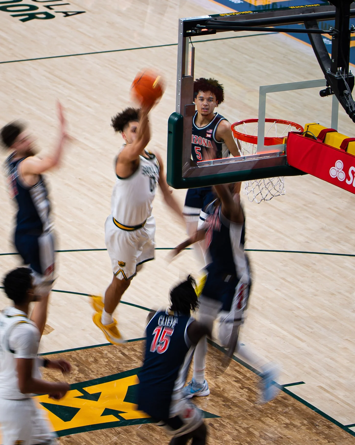 Basketball game with players in action near the hoop, one player in black jersey with 'Arizona' and number 5, jumping to block a shot from a player in white, another player in blue with number 15, on a wooden court.