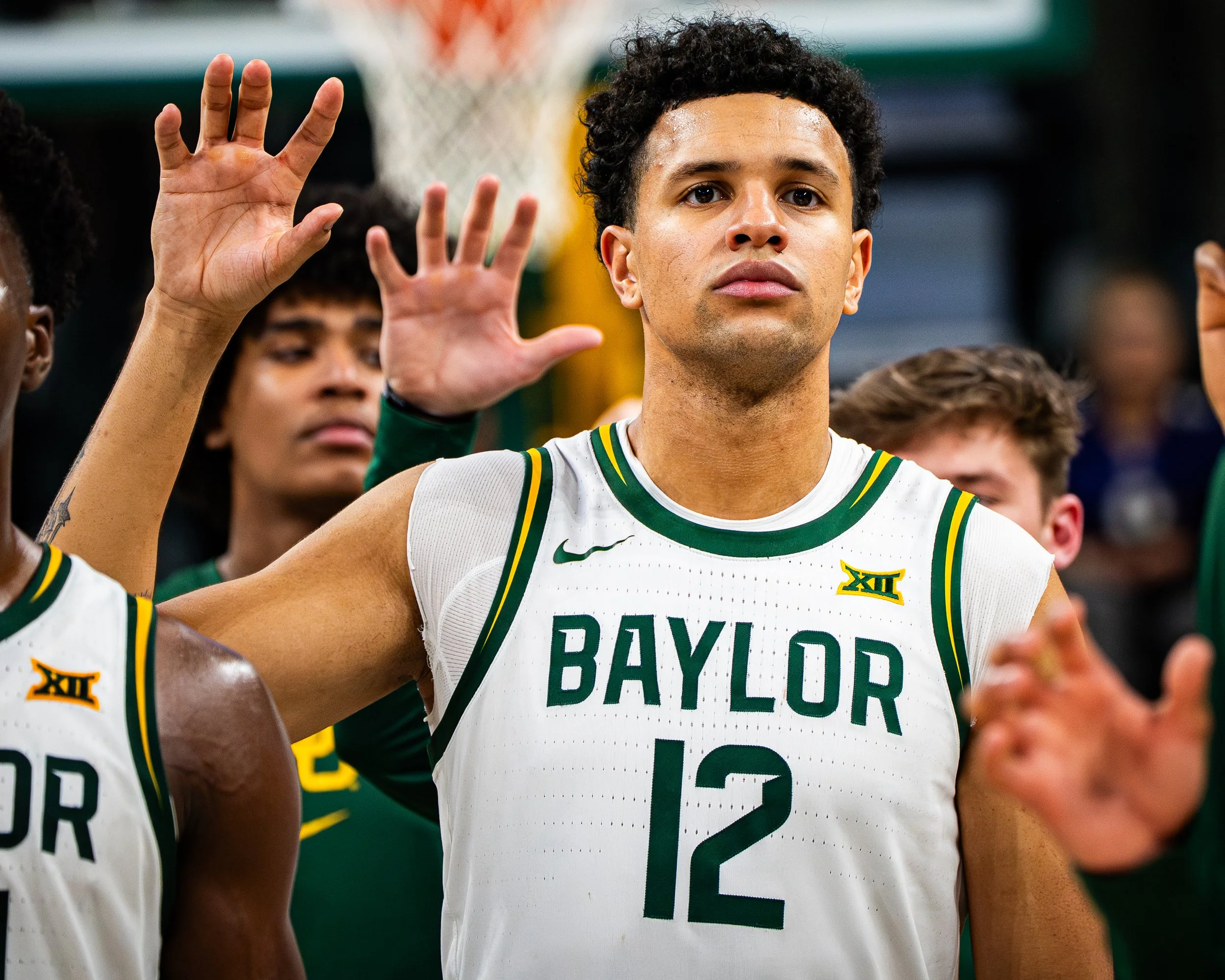 Close-up of a young male basketball player with short black curly hair and sweat on his face, wearing a white Baylor University basketball jersey with green and yellow accents, raising his right hand with three fingers extended during a game or pract