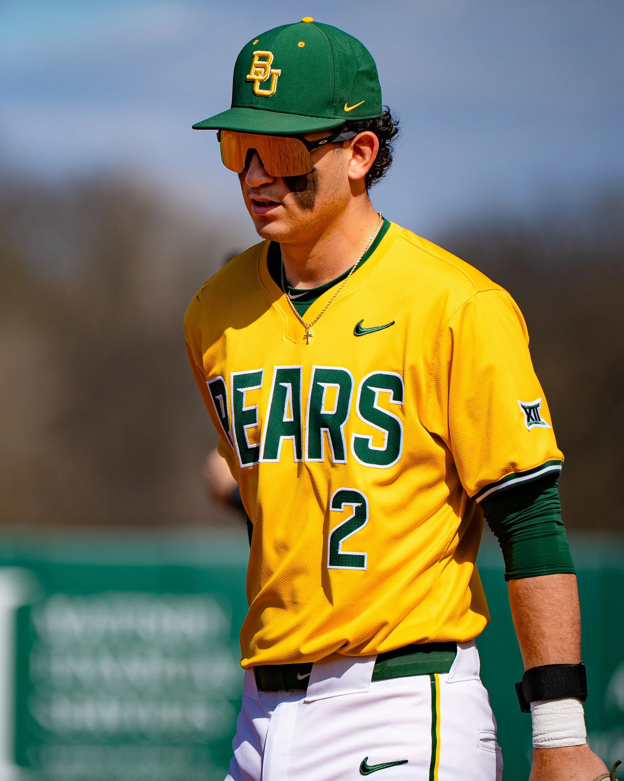 Close-up of a baseball player wearing a yellow jersey, white pants, a green cap with 'BU' logo, reflecting sunglasses, with a background of a baseball field and blue sky.