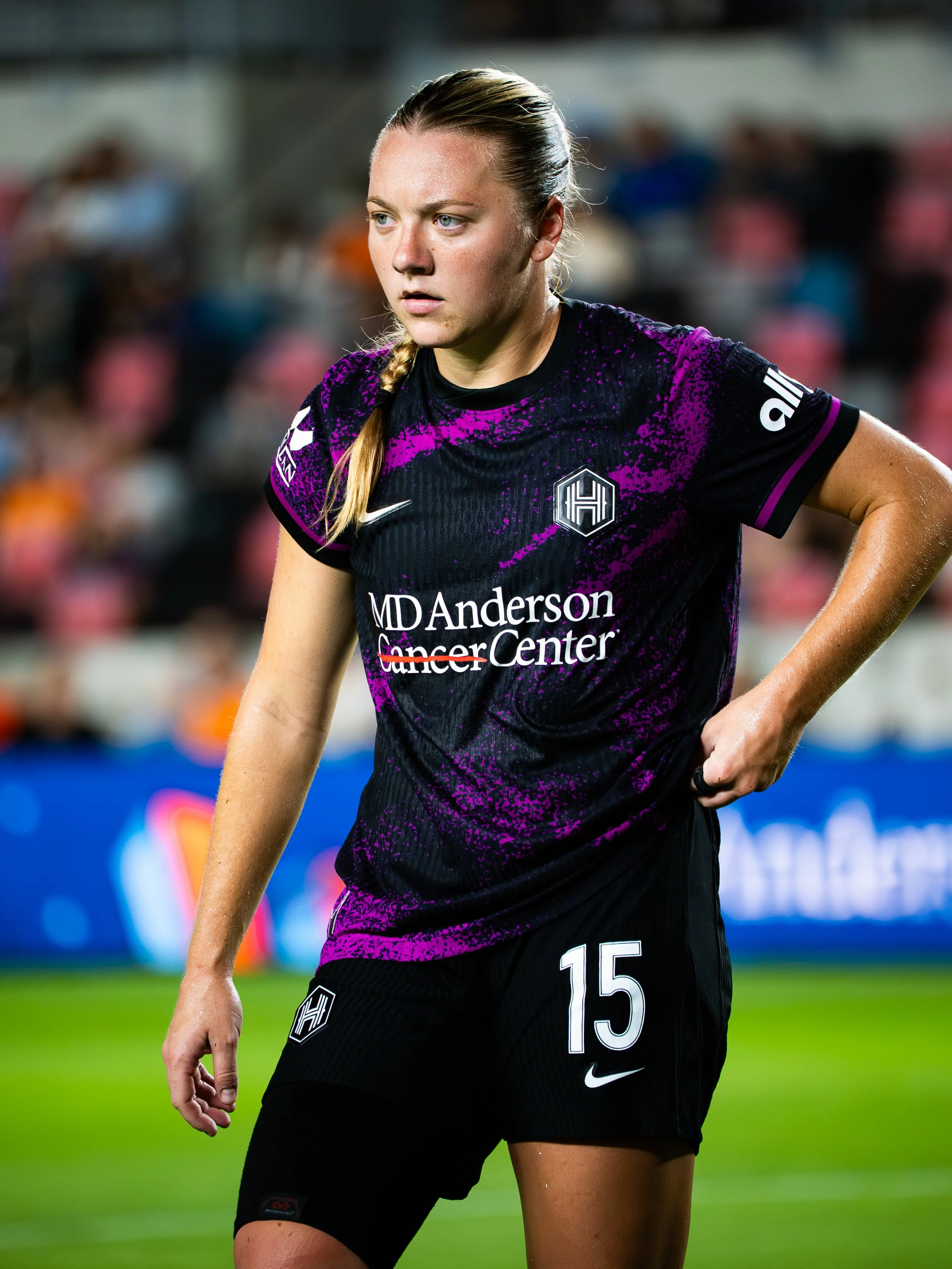 Female soccer player wearing a black and purple uniform with 'MD Anderson Cancer Center' written on it, standing on the field during a game.