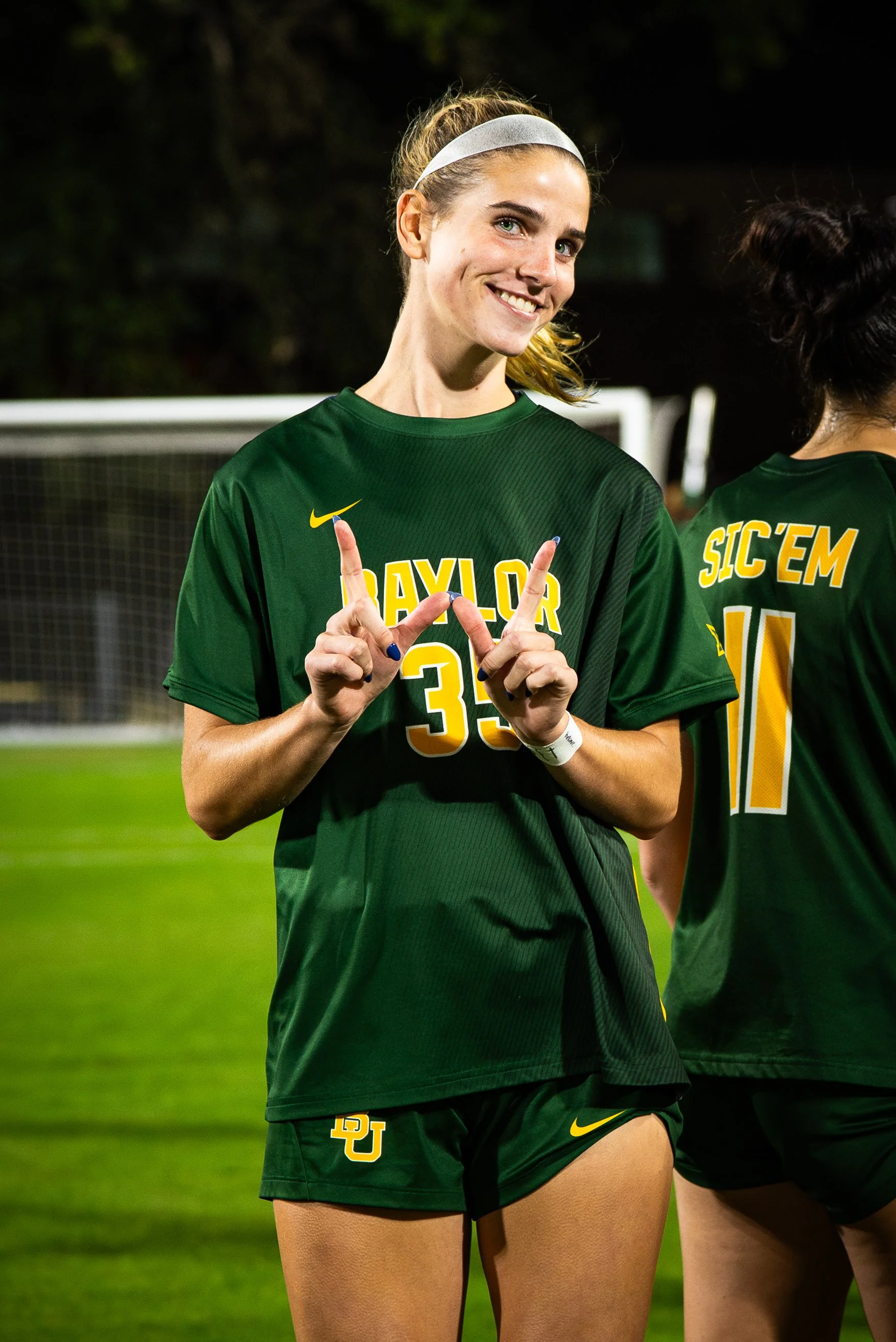 A female soccer player wearing a green uniform with 'BAYLOR' and the number 35, smiling and making a victory sign, standing on a soccer field at night with another player partially visible beside her.