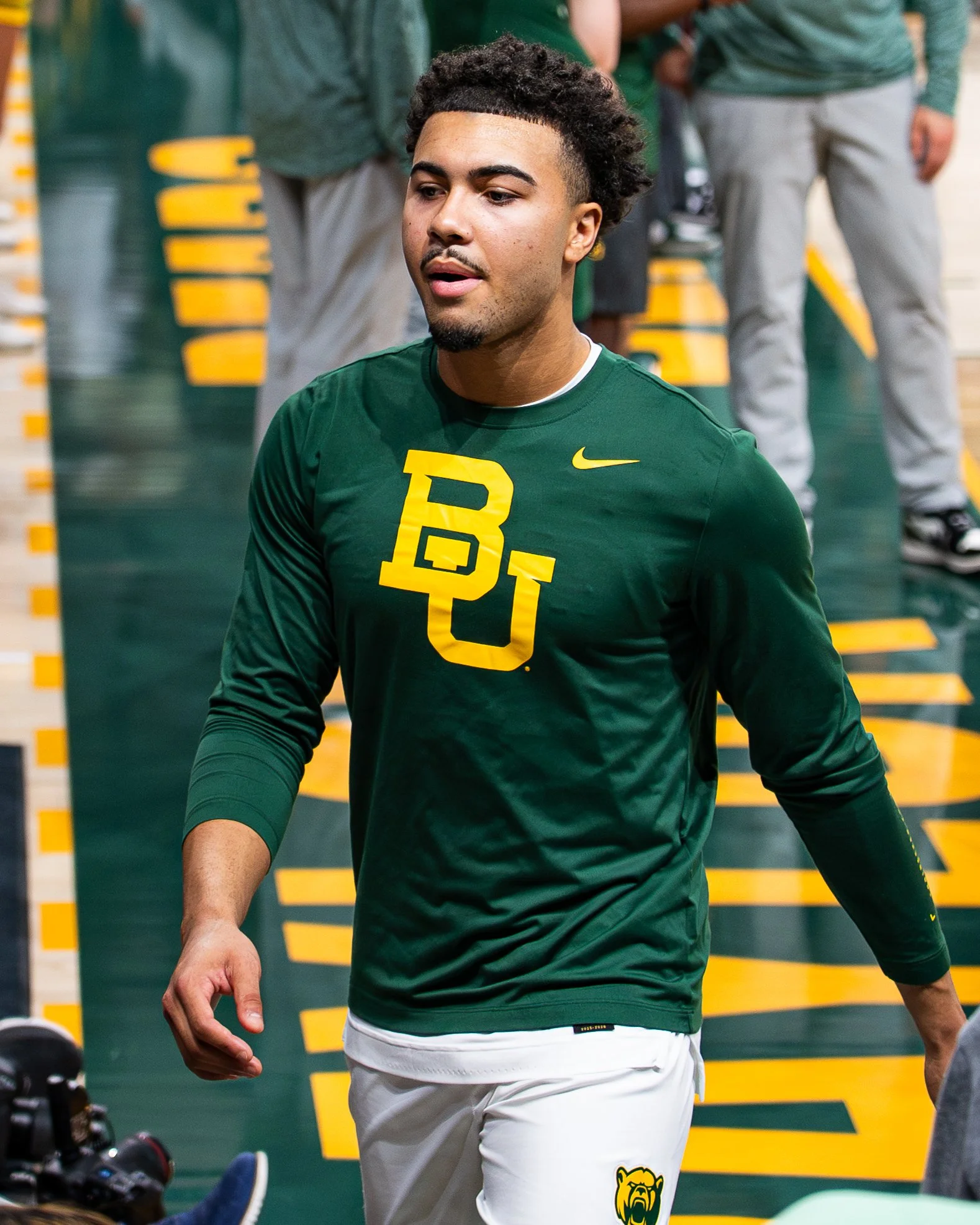 A young man wearing a green long-sleeved shirt with yellow logo and white pants, standing in a sports facility, with a background of people and equipment.