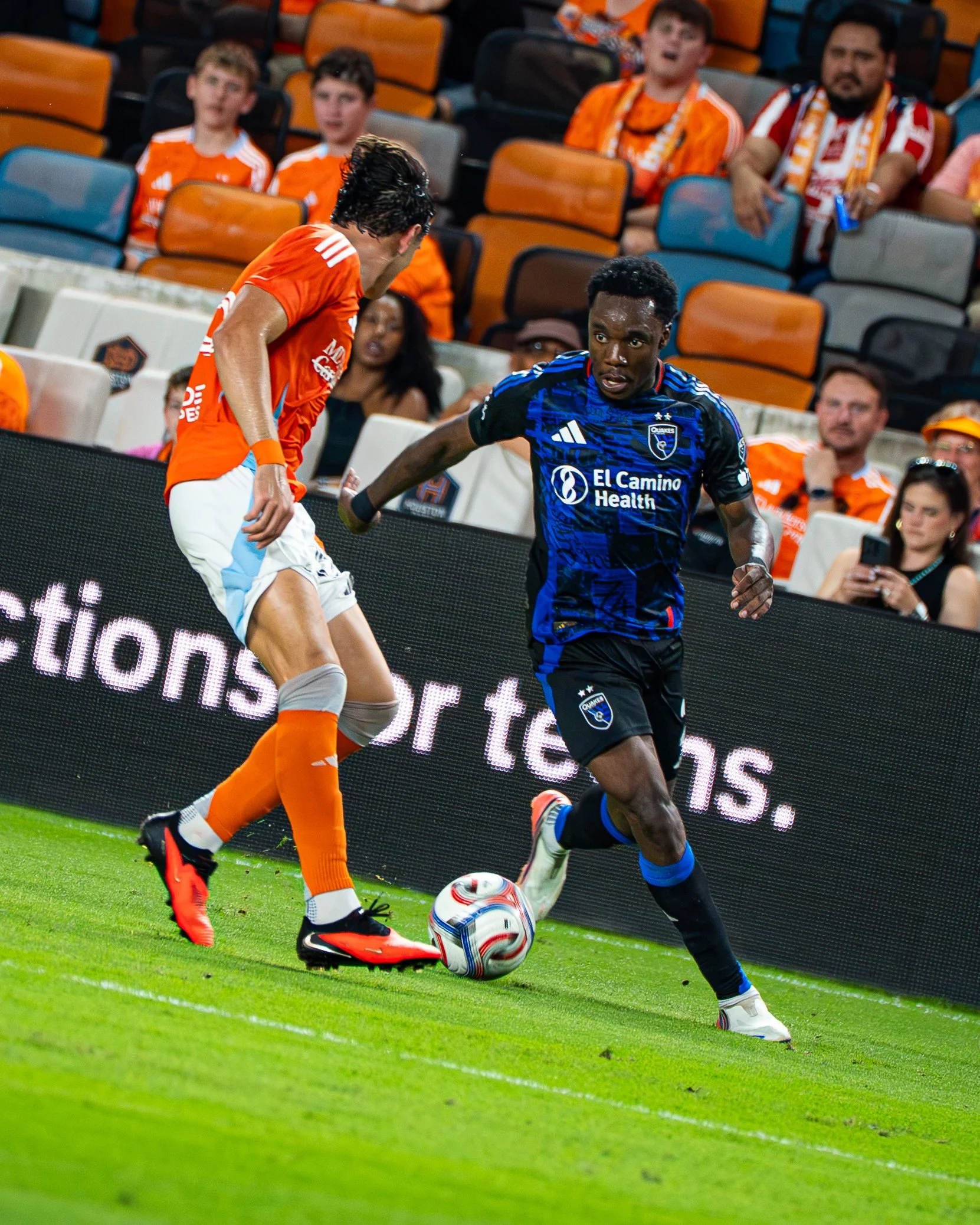 Two soccer players competing for the ball during a match, with spectators watching from the stands.