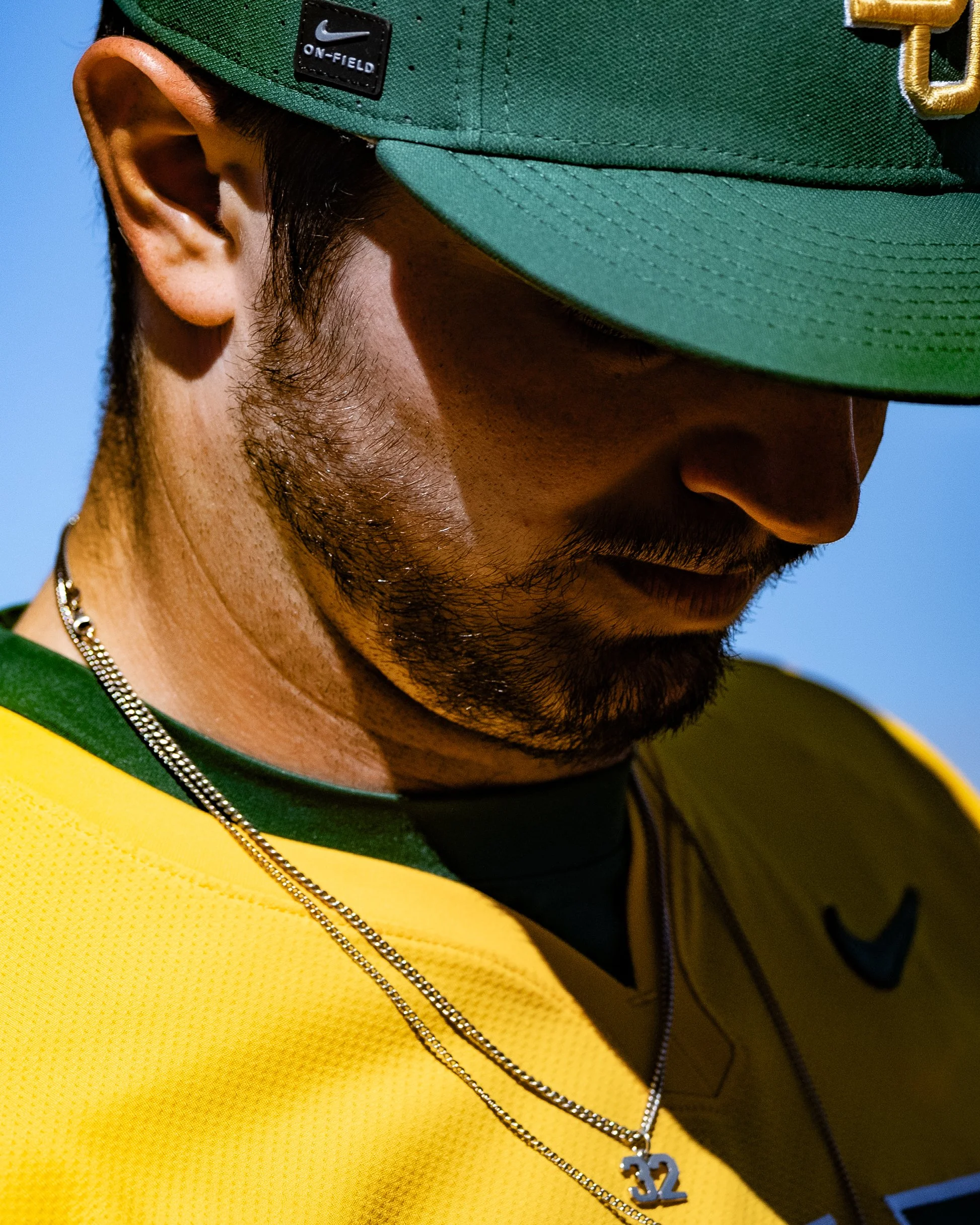 Close-up of a person wearing a green Nike ON-FIELD cap, a yellow sports jersey, and a silver chain necklace with a '32' pendant. The person has facial hair and a small portion of a dark shirt visible beneath the jersey.