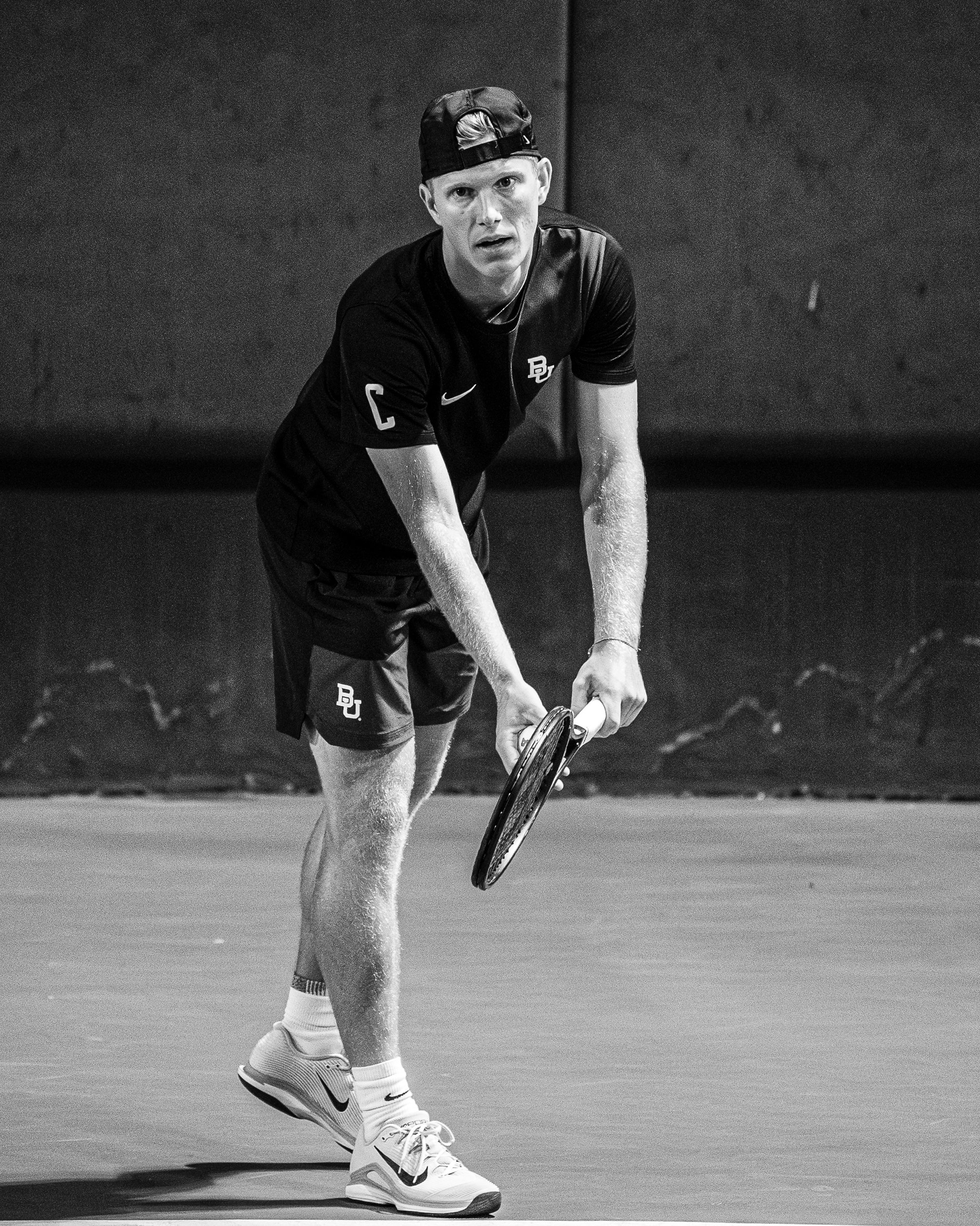 A male tennis player with a backwards cap, wearing a sports shirt with a 'C' and 'BU' logos, shorts, and Nike sneakers, captured mid-action on a tennis court.