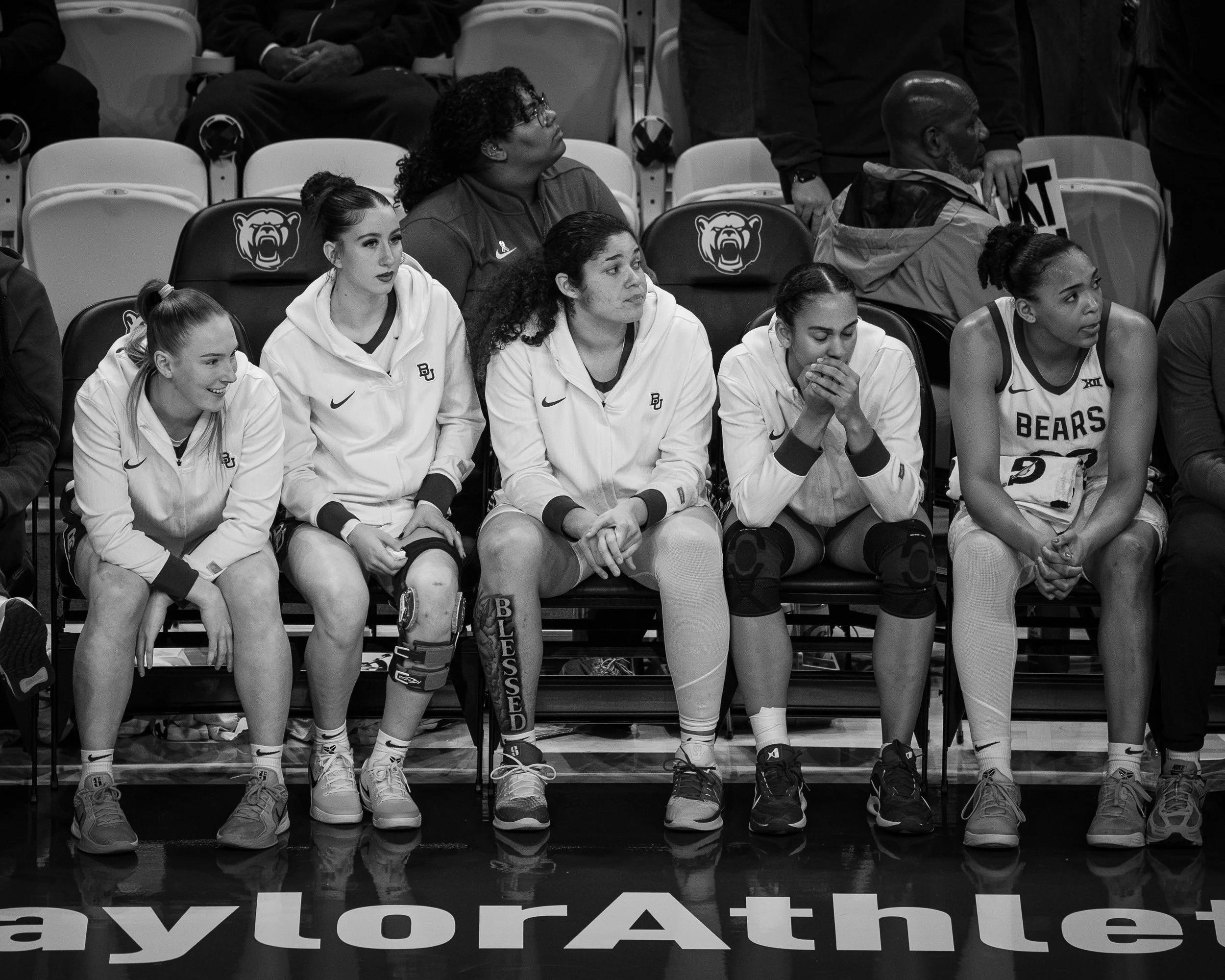 Black and white photo of a group of women athletes sitting on a bench, some wearing white athletic hoodies with 'Baylor' logos, others in sports uniforms. The women appear attentive and focused, with some looking forward and others with hands near th