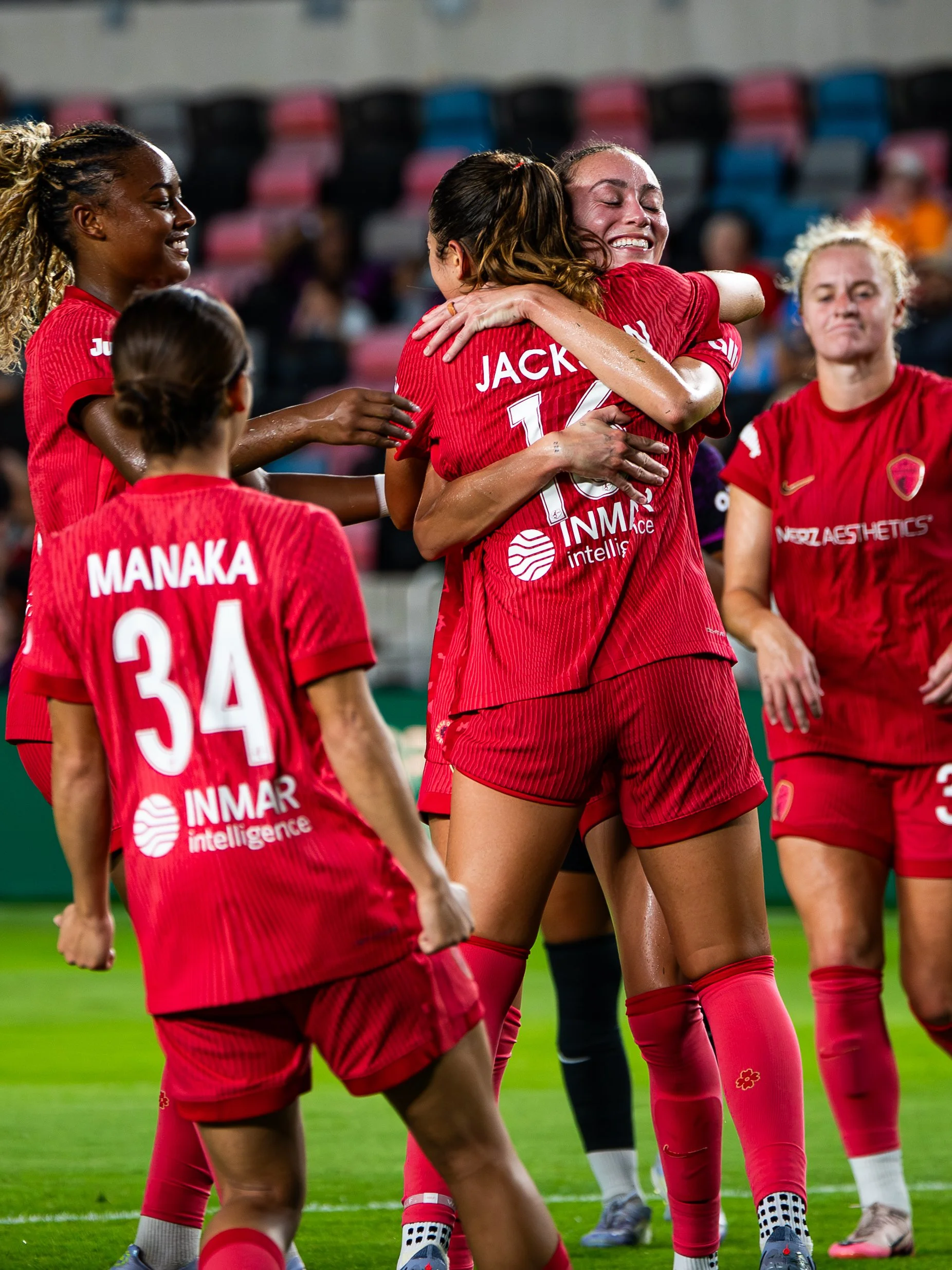 Women soccer players in red uniforms celebrating on the field, hugging each other, with some smiling and some with serious expressions. The background shows rows of stadium seats.