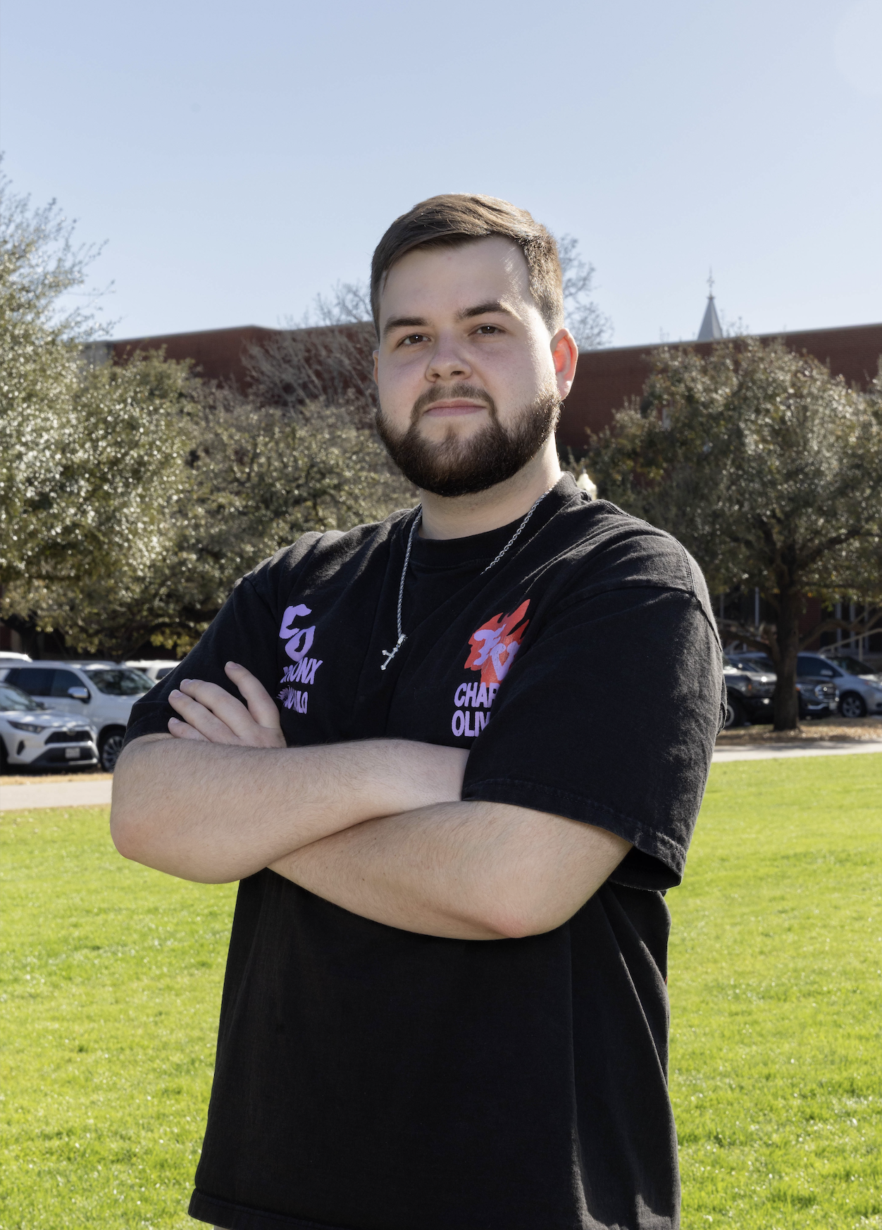A young man with a beard and short hair standing outdoors on a sunny day, arms crossed, with trees, cars, and buildings in the background.