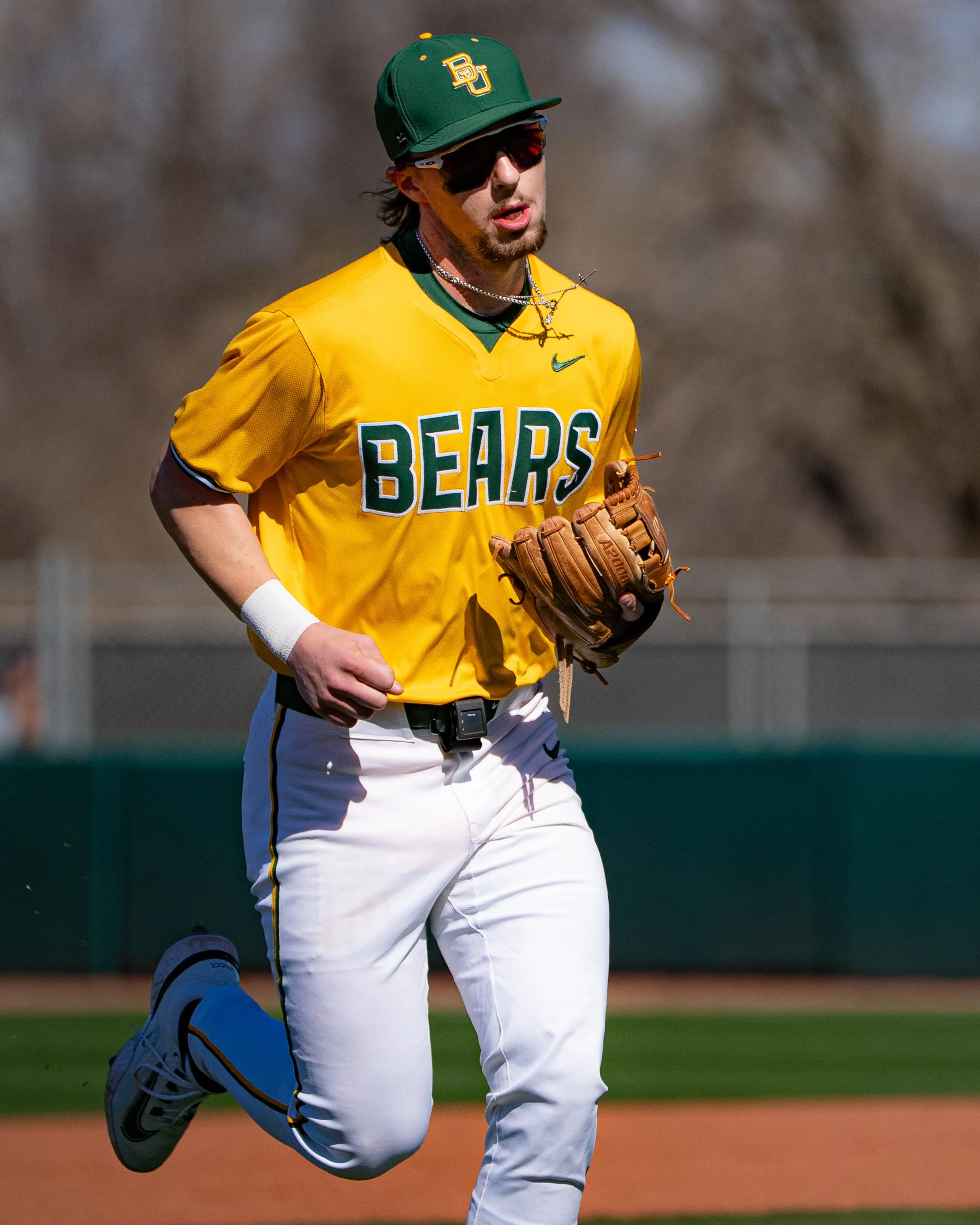 A baseball player wearing a green cap with yellow accents, yellow jersey with 'BEARS' written on it, and white pants with yellow and green stripes is running on the field during daytime. The player is holding a brown baseball glove and wearing sungla
