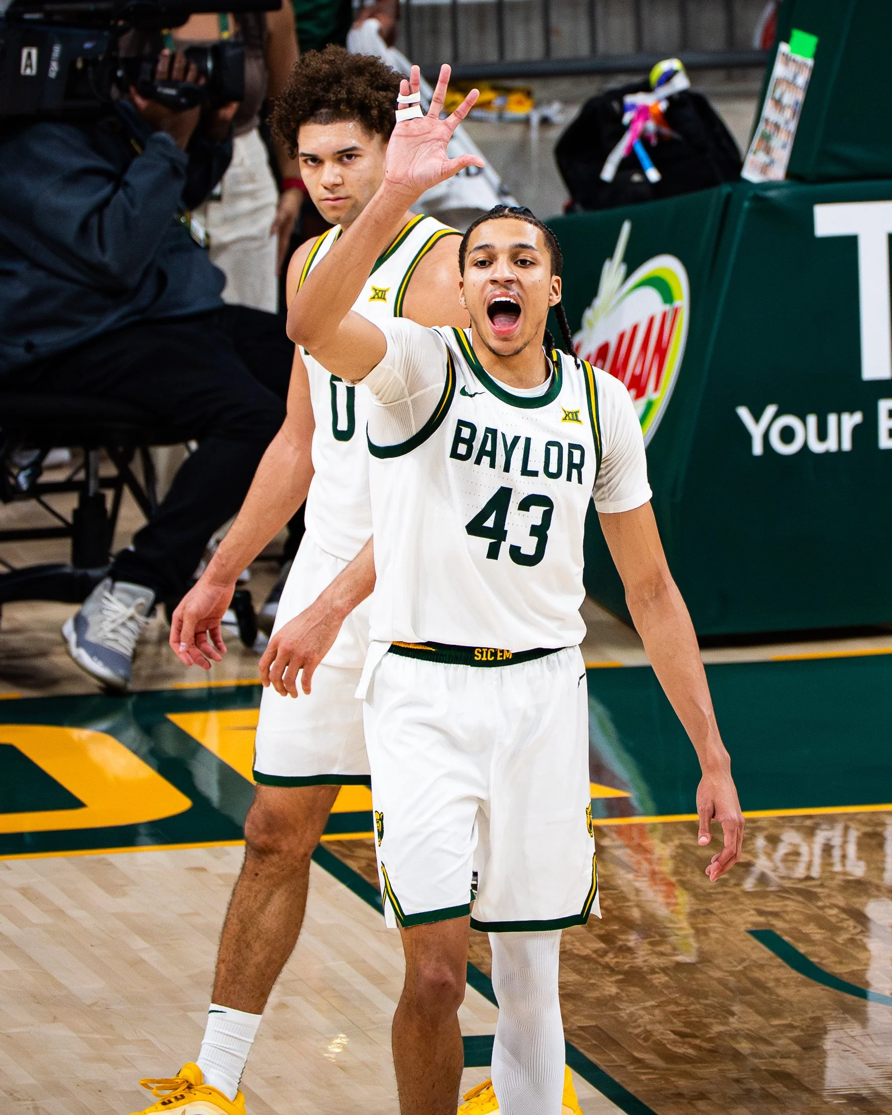 Two basketball players from Baylor University, wearing white jerseys with green and yellow accents, are on the court during a game. One player, with braided hair, is speaking or shouting, and the other player, with curly hair, is raising his hand, po