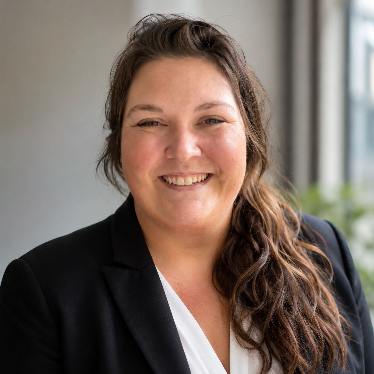 A woman with long, wavy brown hair, smiling, wearing a black blazer and white blouse, in a professional headshot indoors with blurred background and natural light.