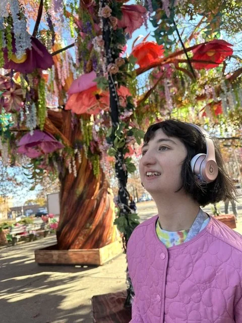 A young girl with short dark hair wearing a pink quilted jacket and silver headphones, standing outdoors near a decorated tree with colorful flowers and hanging ornaments.