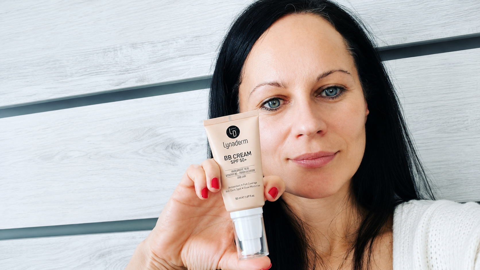 A woman with black hair and blue eyes holding a tube of Lunaderm BB Cream SPF 50+ in front of her face, standing against a white wooden background.