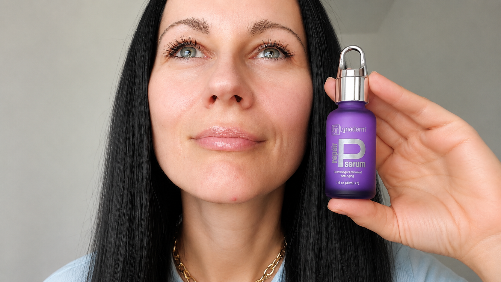 A woman with long black hair and blue eyes holding a purple bottle of serum labeled 'Lynaderm Rejuvenate Serum' close to her face, looking up slightly.