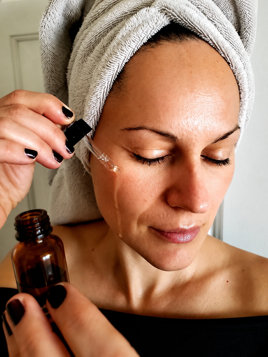 A woman with a towel wrapped around her head applies facial serum from a dropper.