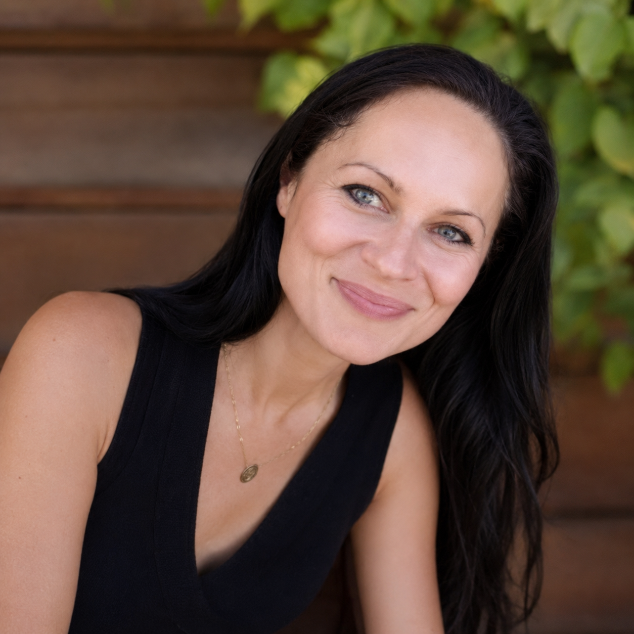 A woman with long black hair and blue eyes smiling outdoors, wearing a black sleeveless top and a gold necklace, with green leaves in the background.