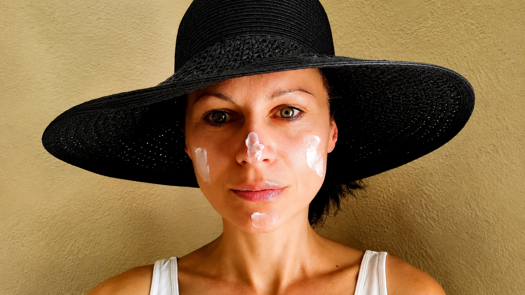 Woman with light skin wearing a large black straw hat, cream-colored tank top, and with white facial cream on her nose and cheeks, standing against a beige wall.