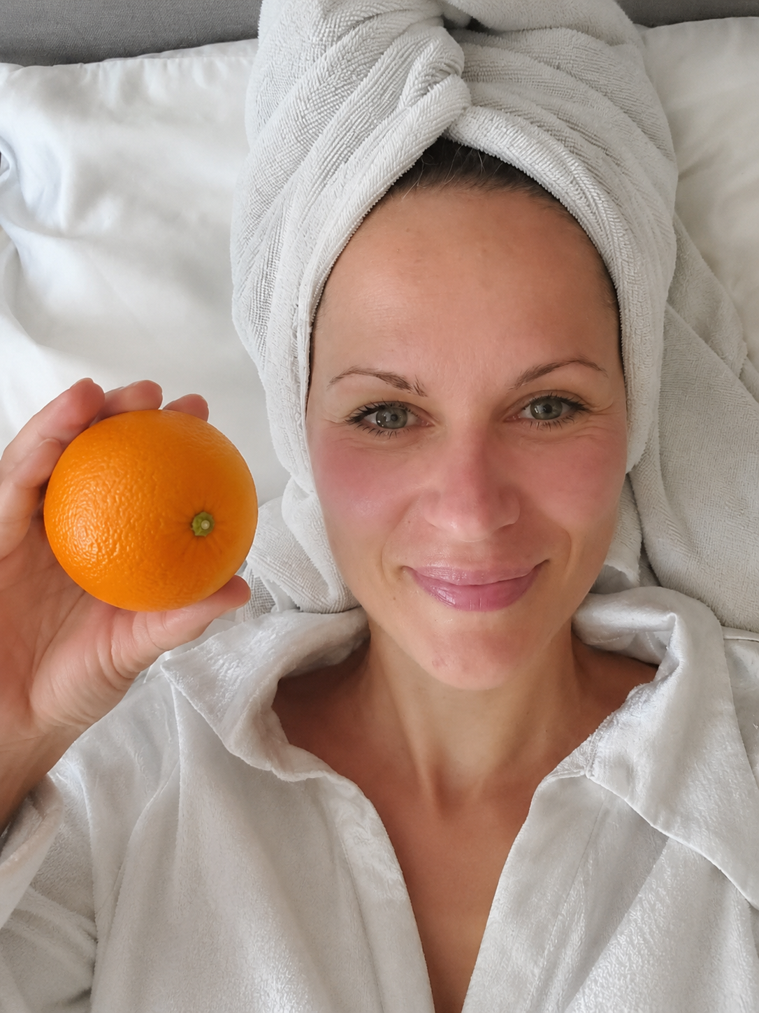 A woman lying in bed, wrapped in a white towel and towel wrapped around head, holding an orange in her left hand, smiling at the camera.