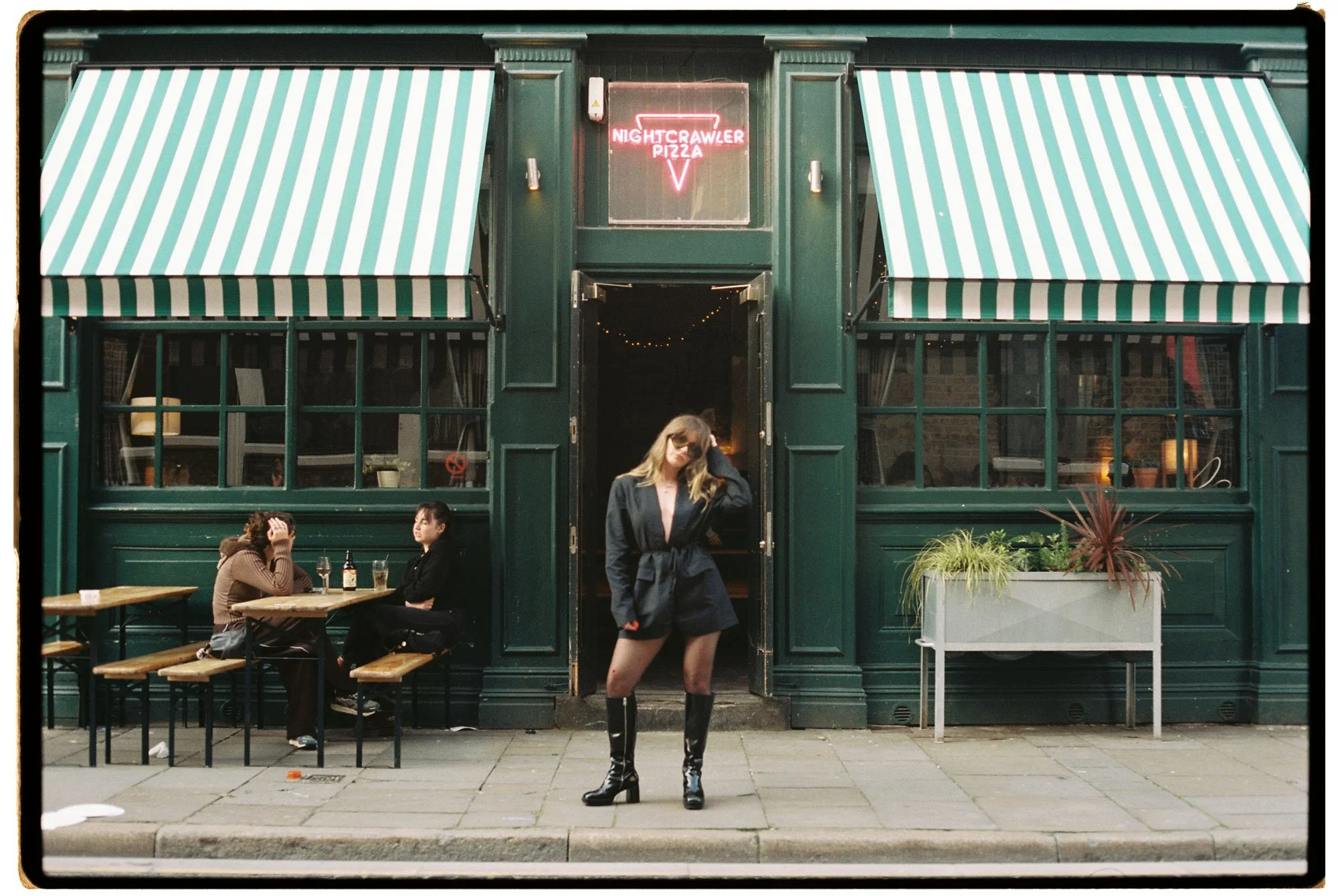 A woman in a black coat and knee-high boots standing on the sidewalk in front of a dark green restaurant with striped awnings. Inside the restaurant, three women are sitting at a table with food and drinks. There is a neon sign above the entrance that reads 'Nightcrawlers Pizza.'