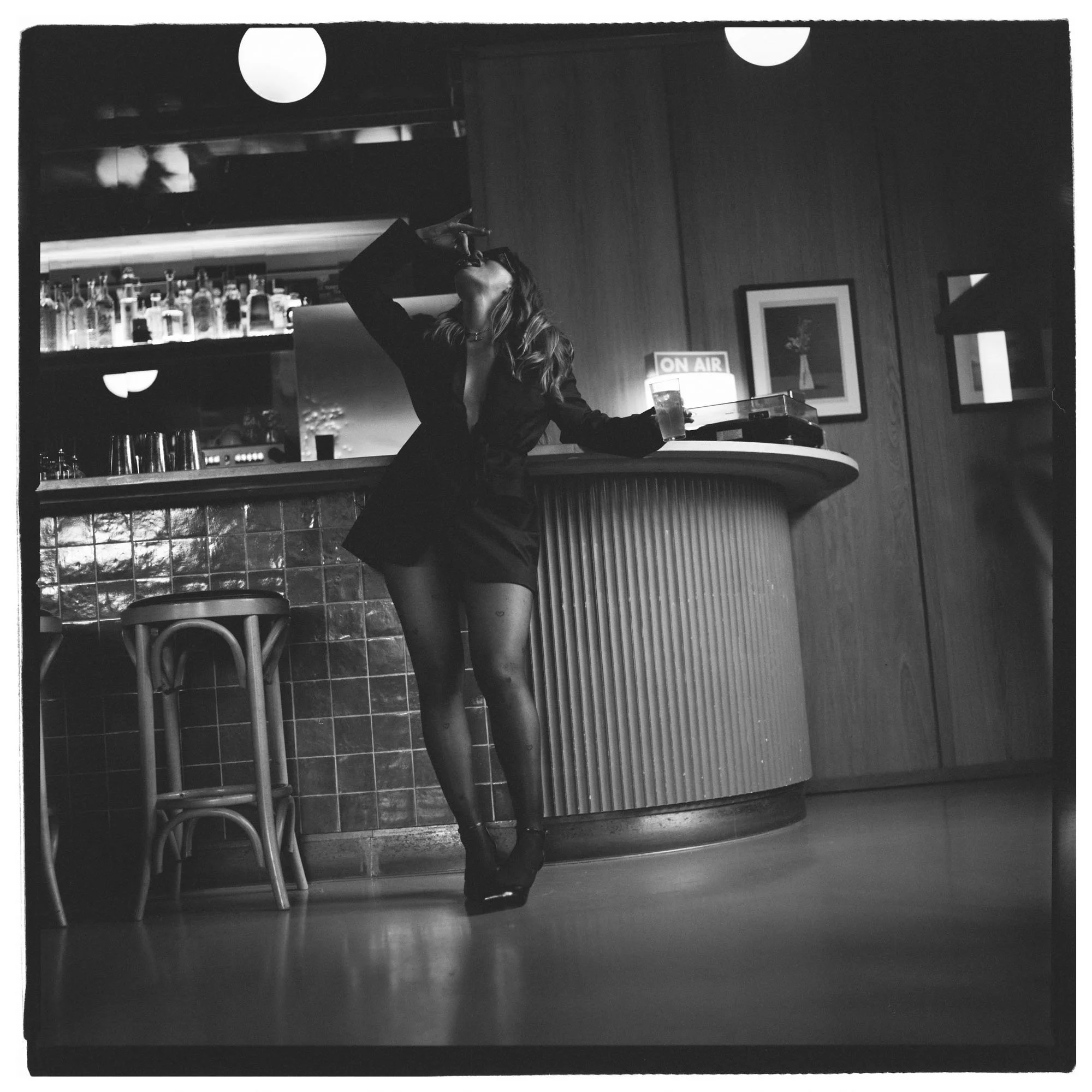 A woman in black clothing, wearing high heels, standing at a bar counter with one hand raised near her face, holding a drink, in a dimly-lit bar or club. There are bottles on shelves in the background and a framed picture on the wall.