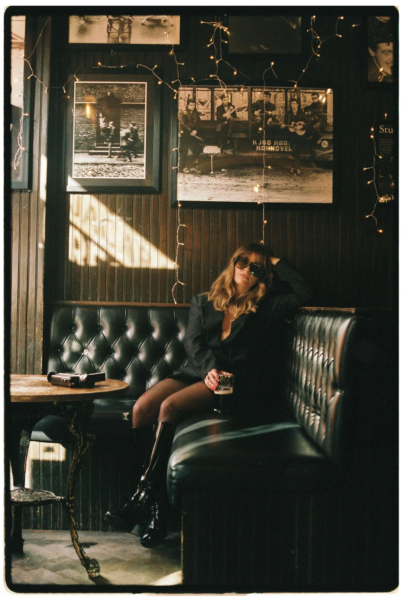 A young woman with wavy hair and sunglasses sits on a black leather booth in a dimly lit bar, holding a beer mug with Guinness beer. She wears a black blazer, patterned tights, and black boots. There are framed black-and-white photos on the wall behind her, along with string lights.