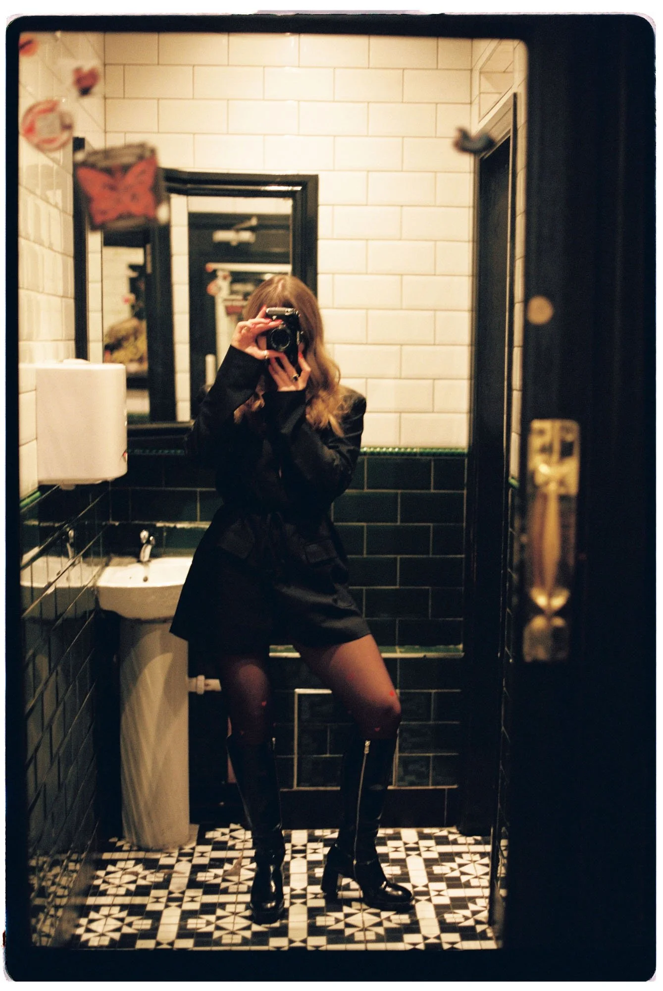 A woman taking a mirror selfie in a bathroom. She is dressed in a black leather jacket, black skirt, sheer tights, and black high-heeled boots. The bathroom has black and white tiled floors, beige brick walls, a sink, a paper towel dispenser, and a mirror with multiple reflections.