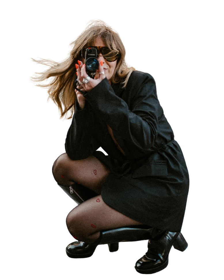 Woman taking a photo with a camera, wearing sunglasses, black jacket, black skirt, and black boots, with small red heart drawings on her legs.
