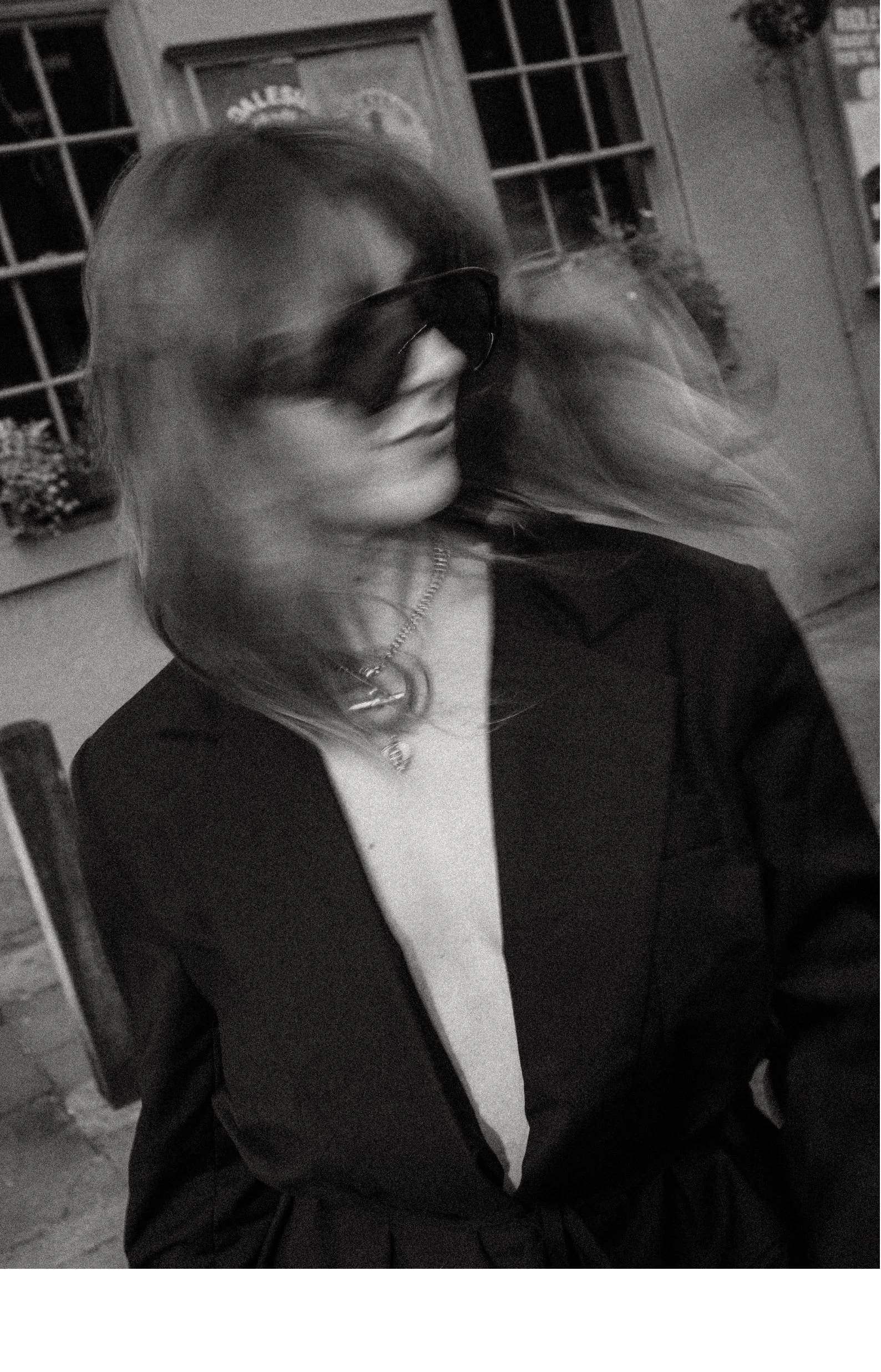 A woman wearing sunglasses, a necklace, and a blazer, standing indoors near a window and a brick wall.