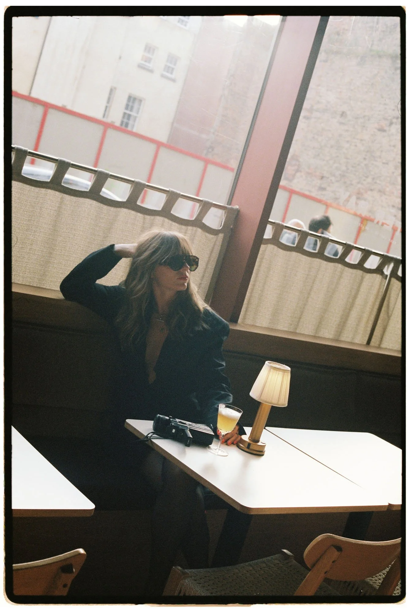 A woman with long dark hair and sunglasses, sitting in a cafe with a drink, a camera, and a lamp on the table. Large windows show buildings outside.