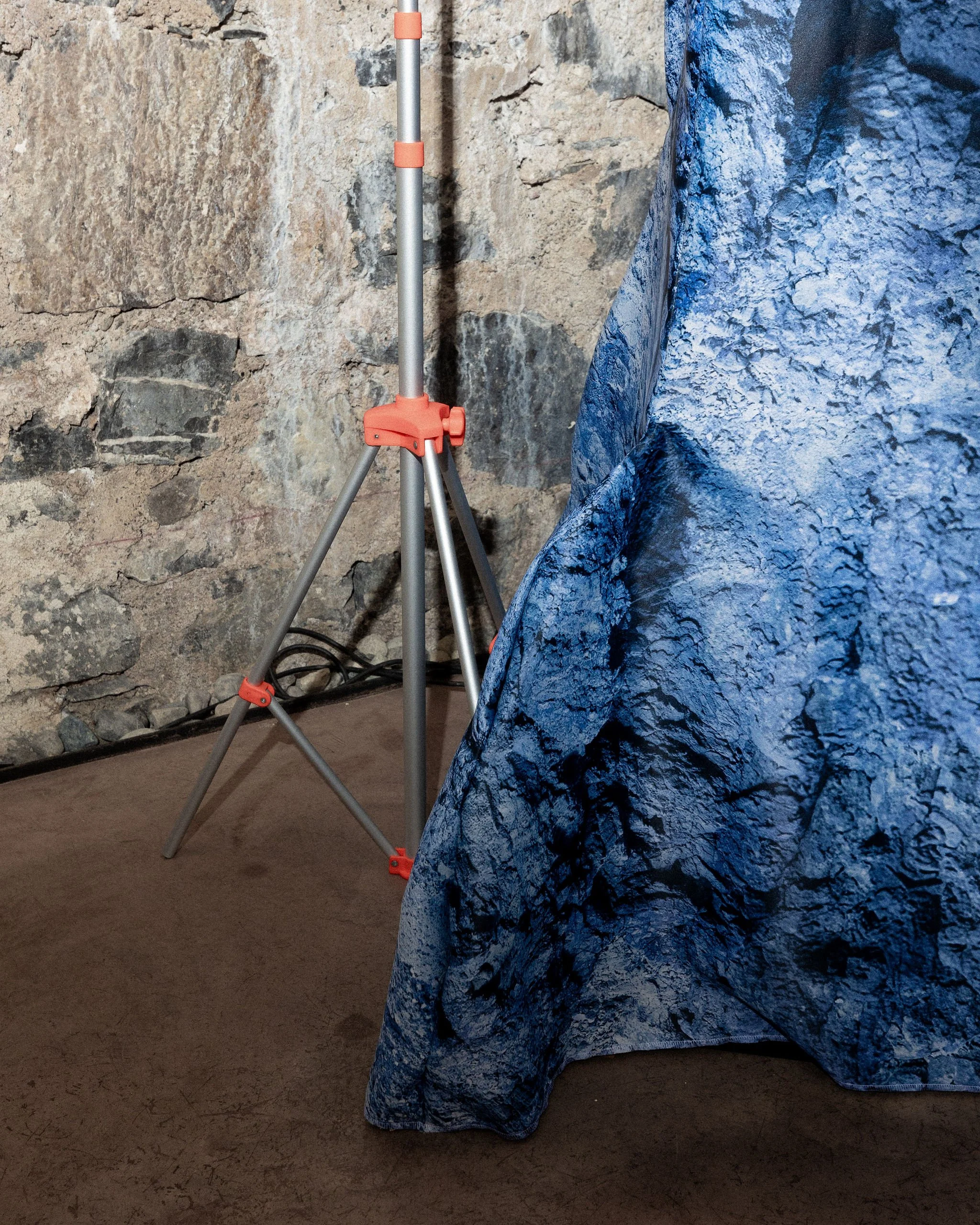 Une scène en intérieur avec un mur en pierre, une toile bleue et un trépied orange et gris. La toile semble représenter une roche ou un paysage rocheux.