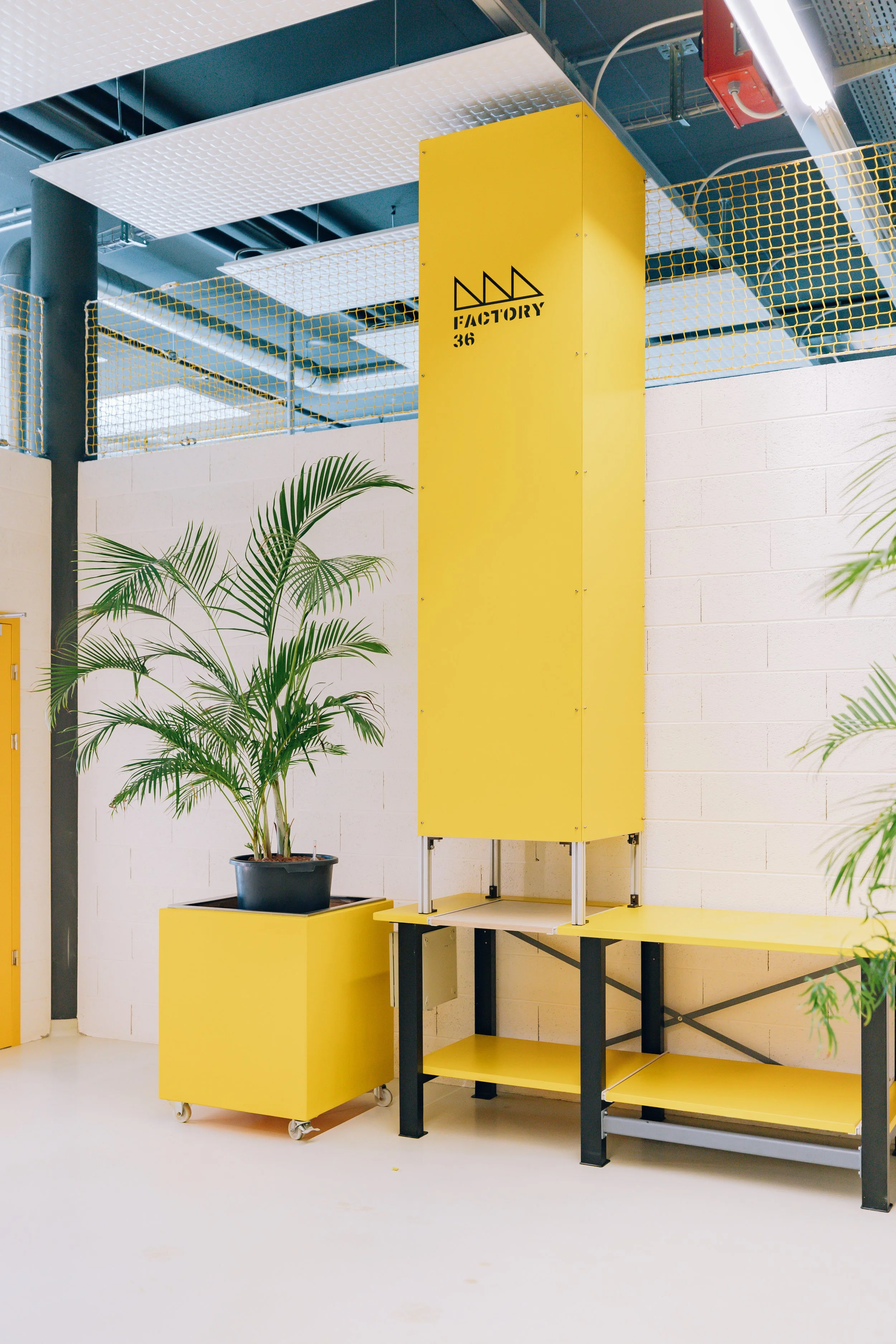 Espace intérieur moderne avec un panneau jaune portant le texte 'FACTORY 36', un étal de travail noir et jaune, des plantes vertes en pot, et un plafonnier industriel.