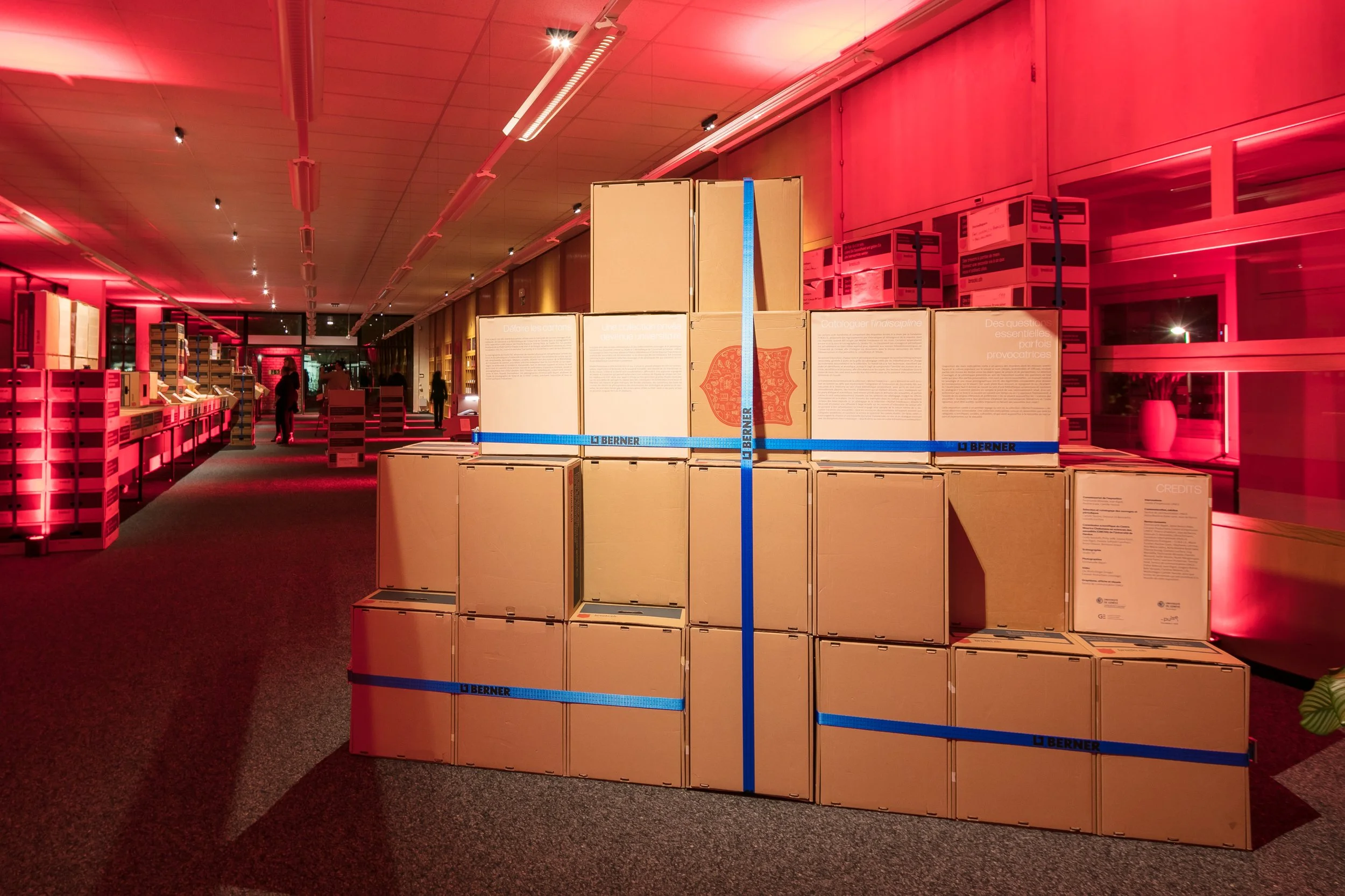 Une pile de cartons composent une scénographie dans une exposition sur la sexualité, emballés avec des sangles bleues. Sur les cartons on trouve le texte de présentation de l'exposition en français avec quelques personnes et étagères en arrière-plan.