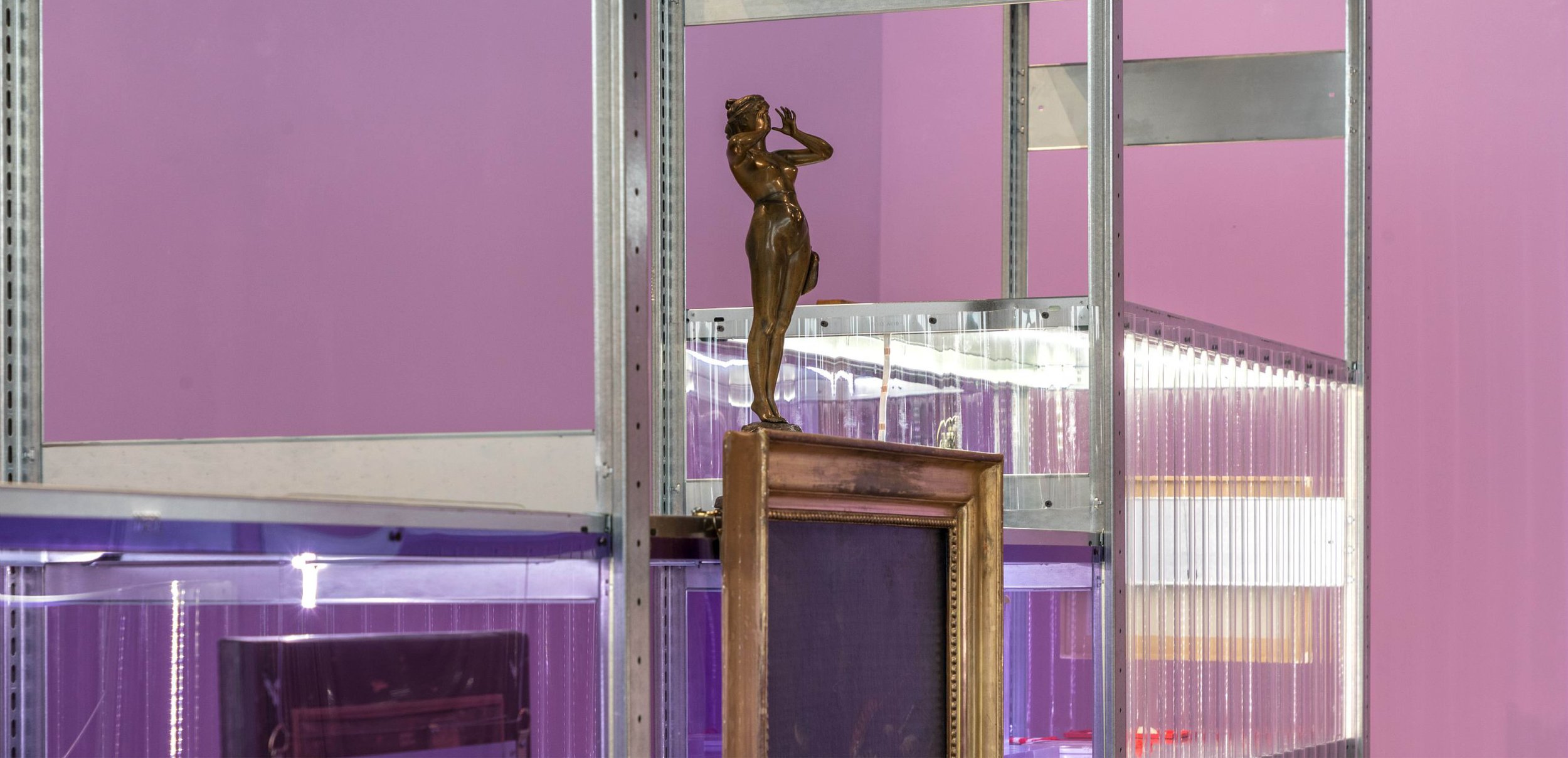Une sculpture en bronze d'une femme en position pensive, placée sur un socle en bois, visible à travers une structure métallique dans une pièce aux murs violets.