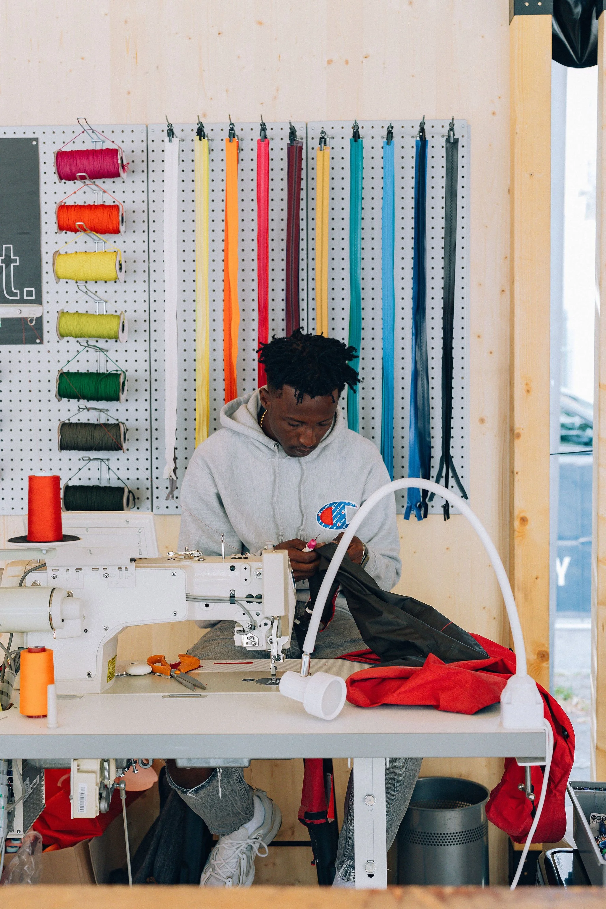 Une personne en train de coudre à une machine à coudre dans un atelier, avec des bobines de fil coloré suspendues au mur derrière elle.