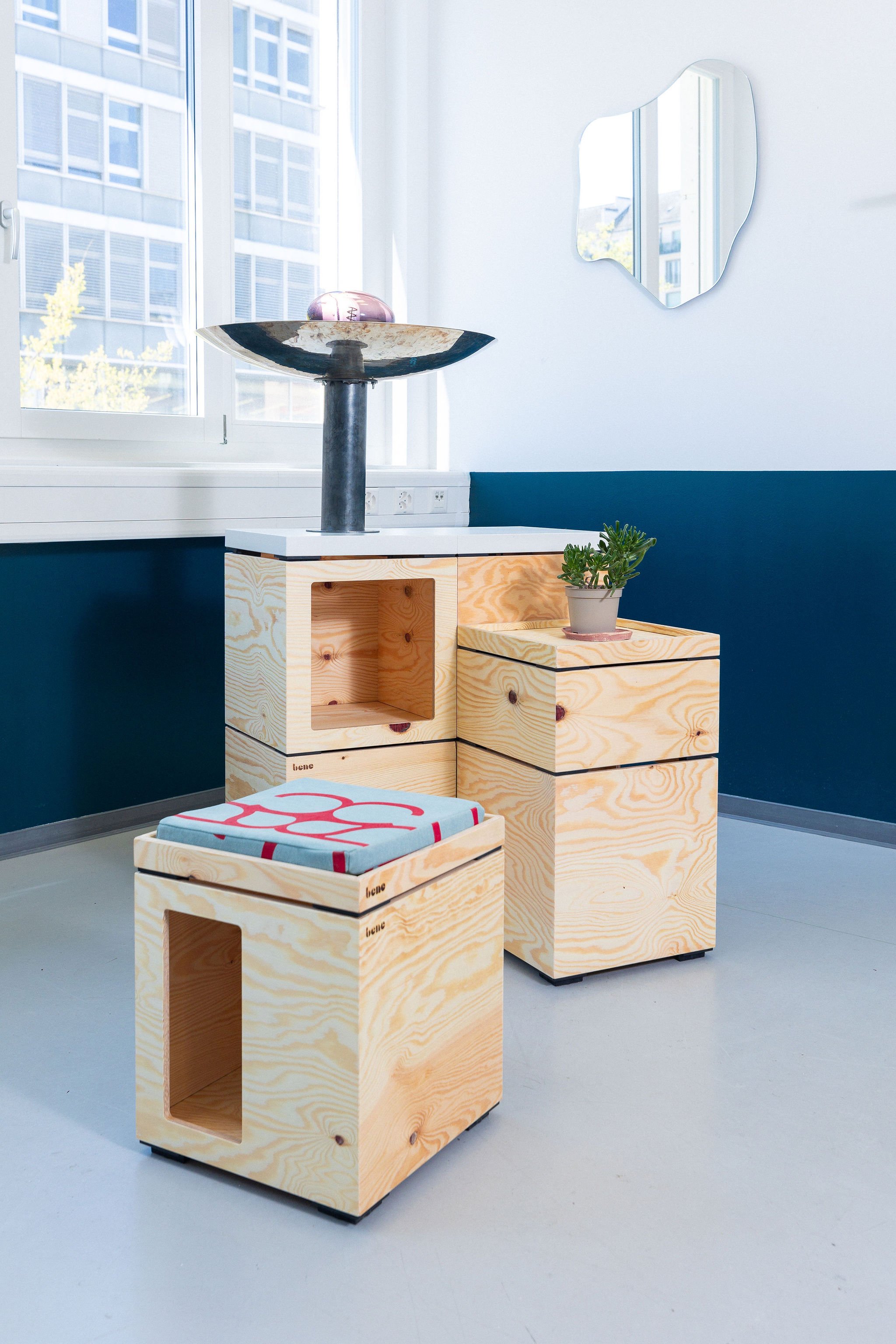Mobilier en bois clair composé de plusieurs caisses empilées, dont une sert de siège avec un coussin bleu et rouge, une autre supporte une plante en pot, et une autre une sculpture ou un objet artistique, dans une pièce avec mur blanc et mur bleu foncé, et une fenêtre laissant entrer de la lumière naturelle.