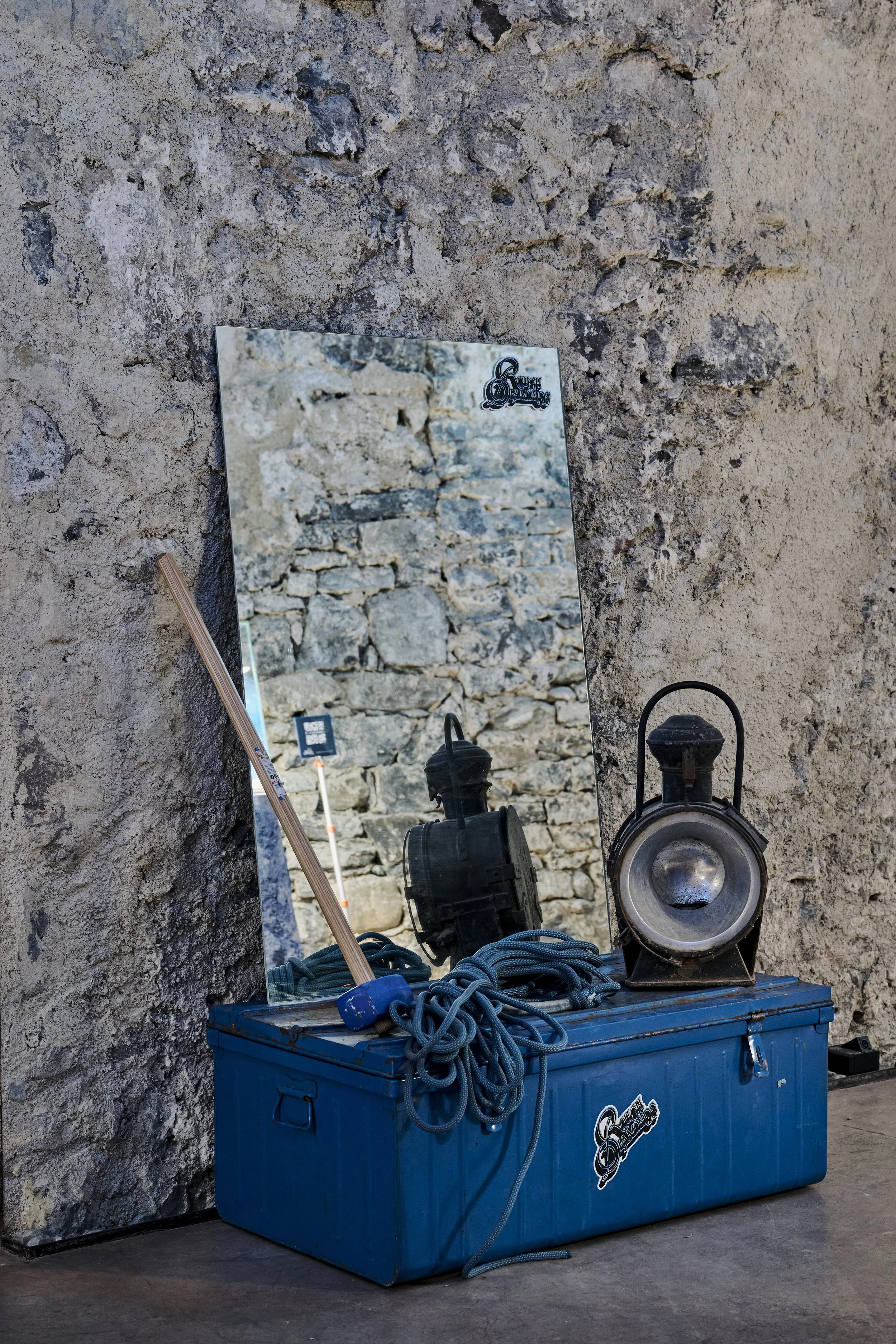 Un miroir posé contre un mur en pierre reflète un fond également constitué de pierres. Devant le miroir, une boîte en métal bleu avec des câbles, un projecteur et une brosse en bois sont posés, avec une étiquette ou un autocollant "Slow Dream".