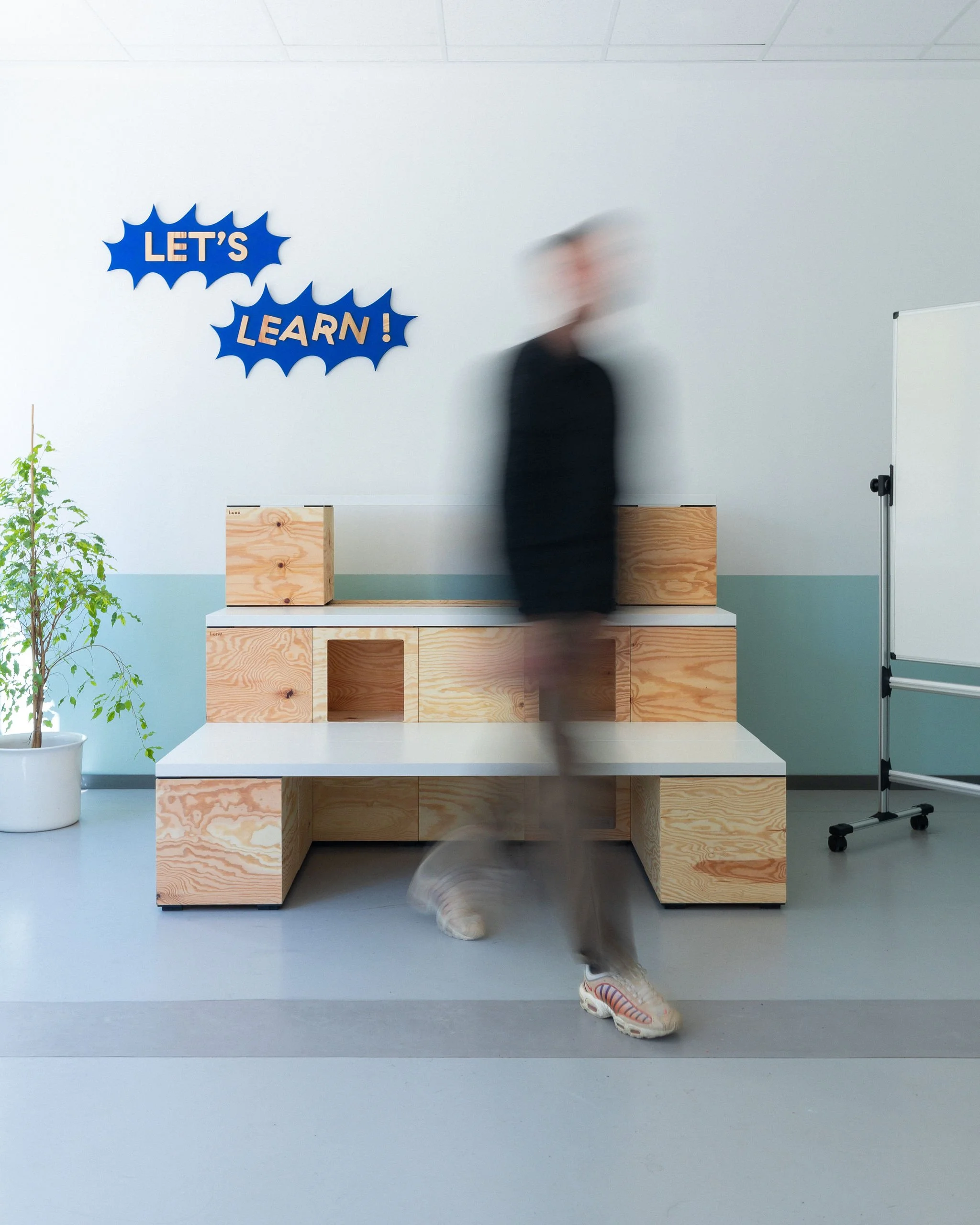 Une salle de workshop dans un coworking avec du mobilier modulable en bois clair, un bureau et étagères, des bancs, deux plantes, un mur blanc avec une décoration en forme de nuages bleus avec des inscriptions en français « LET'S LEARN ! ».