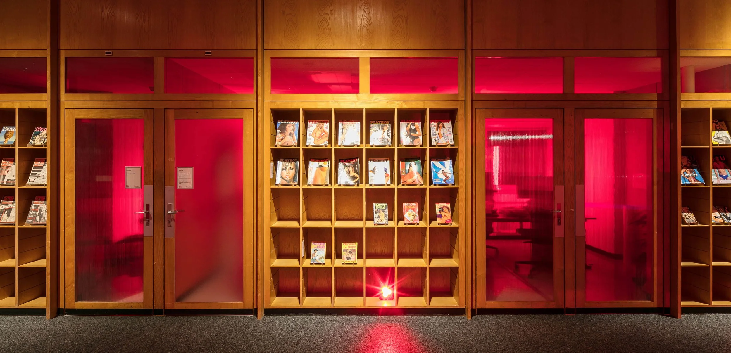 Une rangée de magazines de magazines pour adultes sur des étagères en bois derrière des portes en verre rouge.
