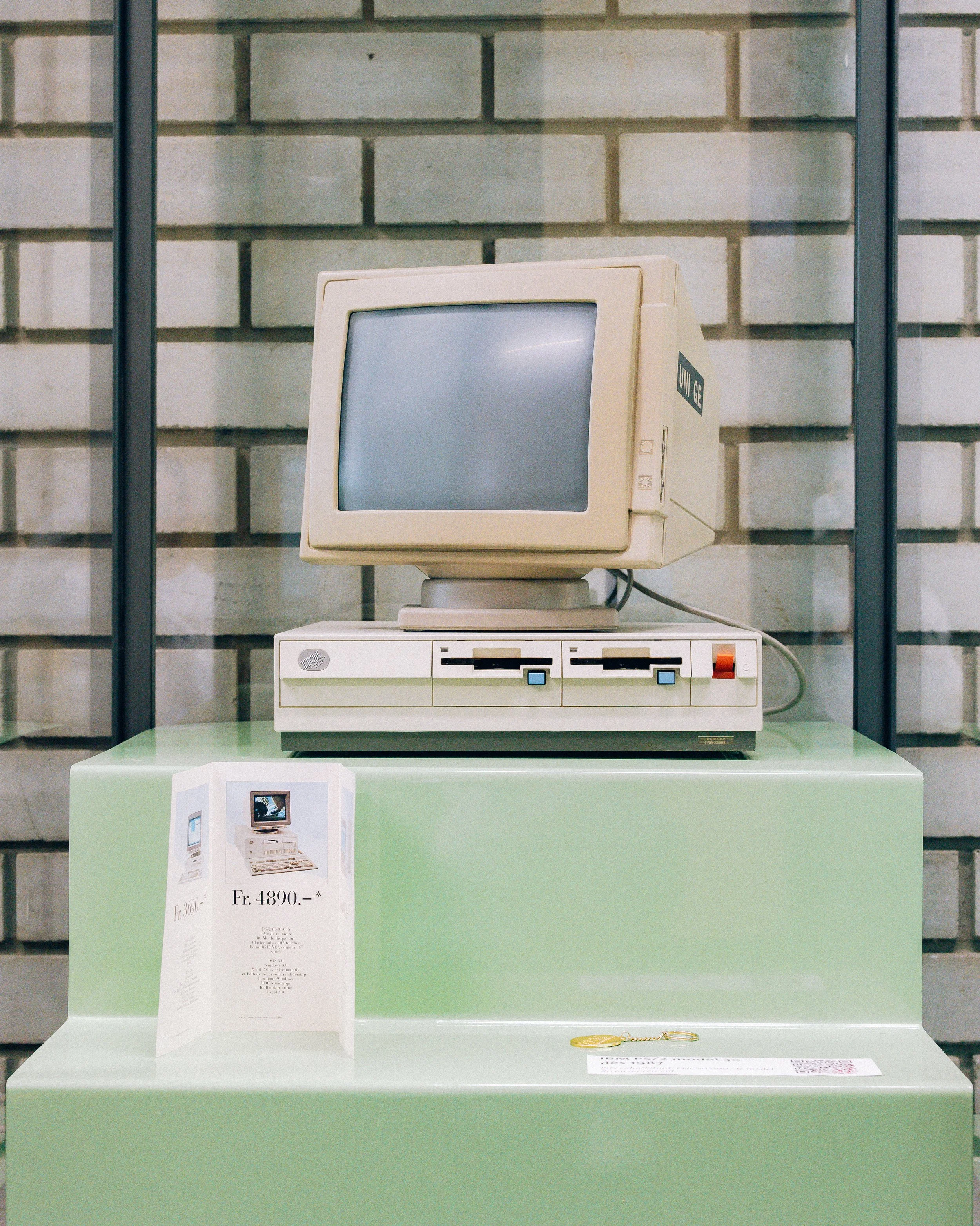 Une vitrine contenant un ancien ordinateur avec un écran beige et une unité centrale, placé sur une table verte. Un poster avec le prix fr. 4890.- et des détails est placé devant l'ordinateur, avec une clé à côté. La vitrine a des portes en verre et un mur de briques en arrière-plan.