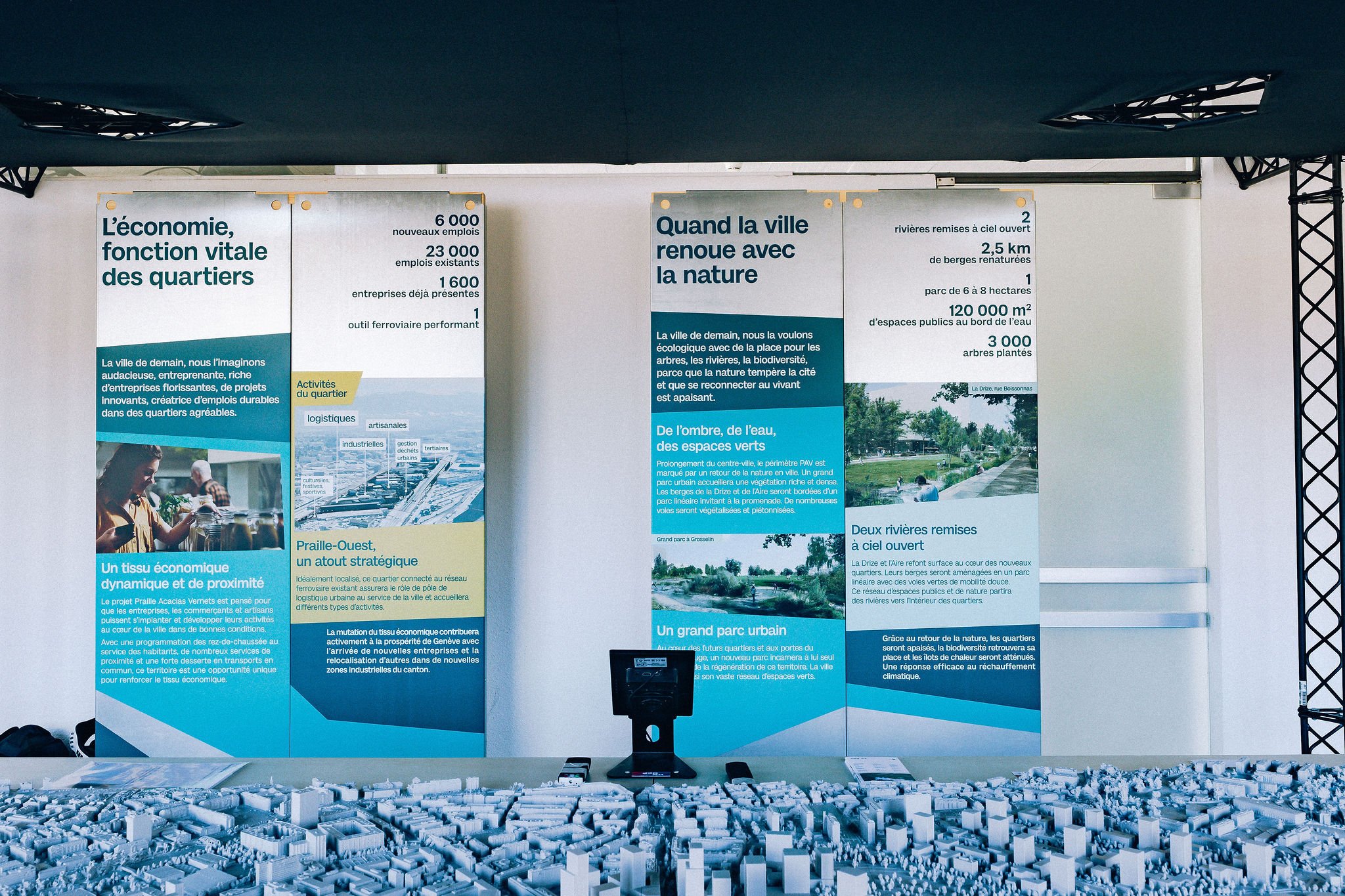 Scénographie d'exposition du projet urbanistique Praille Acacias Vernet à Genève. Affiches informatives sur la revitalisation urbaine et la nature dans les quartiers, avec images de parc et de bâtiments, et expositions en français.